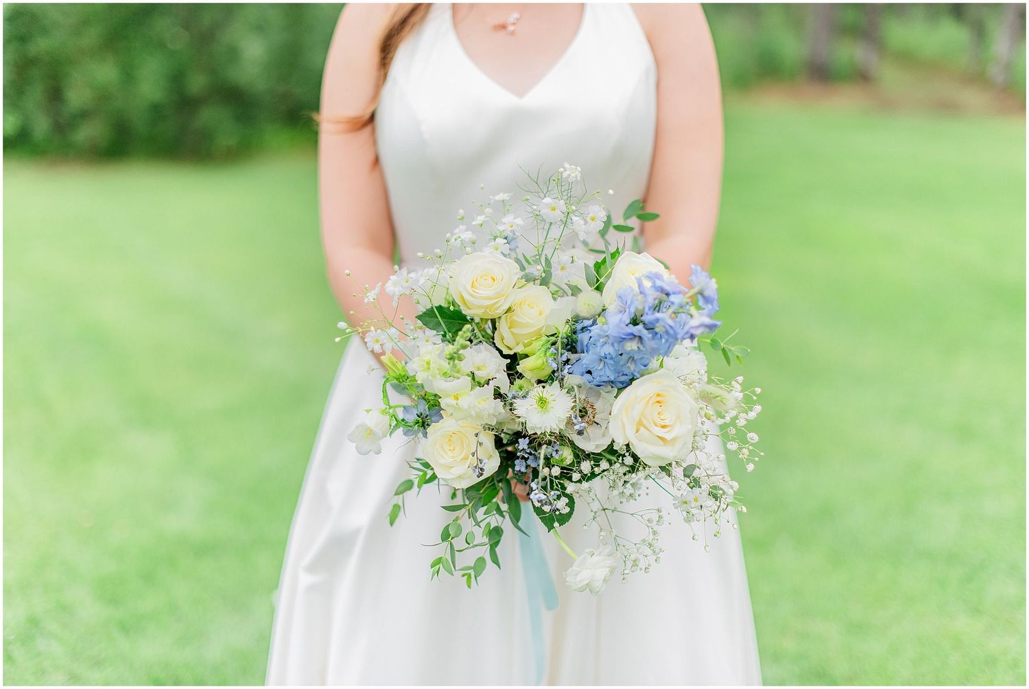 Bride holding a stunning bouquet of white roses, blue hyacinths, and greenery in a white satin wedding gown.