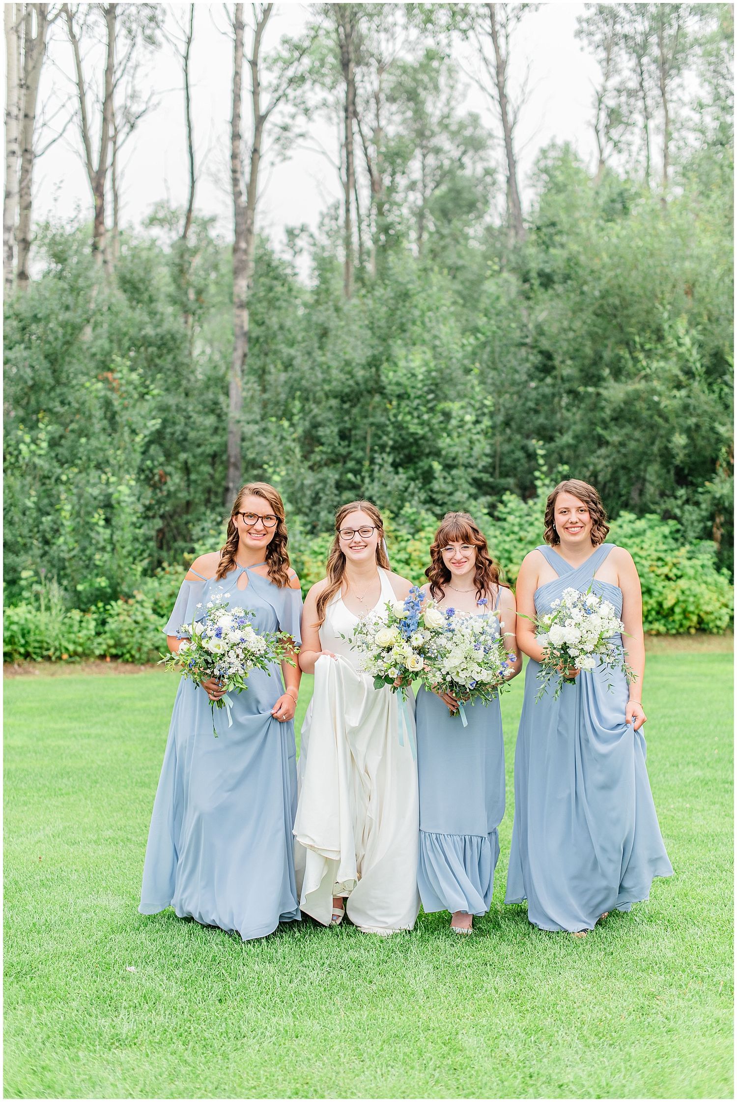 Bride in ivory gown poses with three bridesmaids in dusty blue dresses holding wildflower bouquets outdoors.