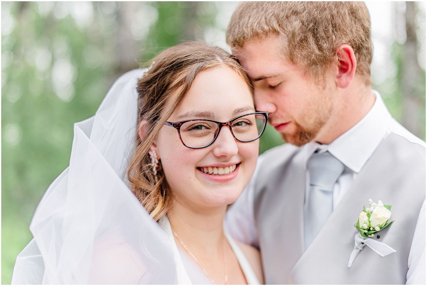 Smiling bride with glasses in white veil poses with groom in gray suit with floral boutonniere.
