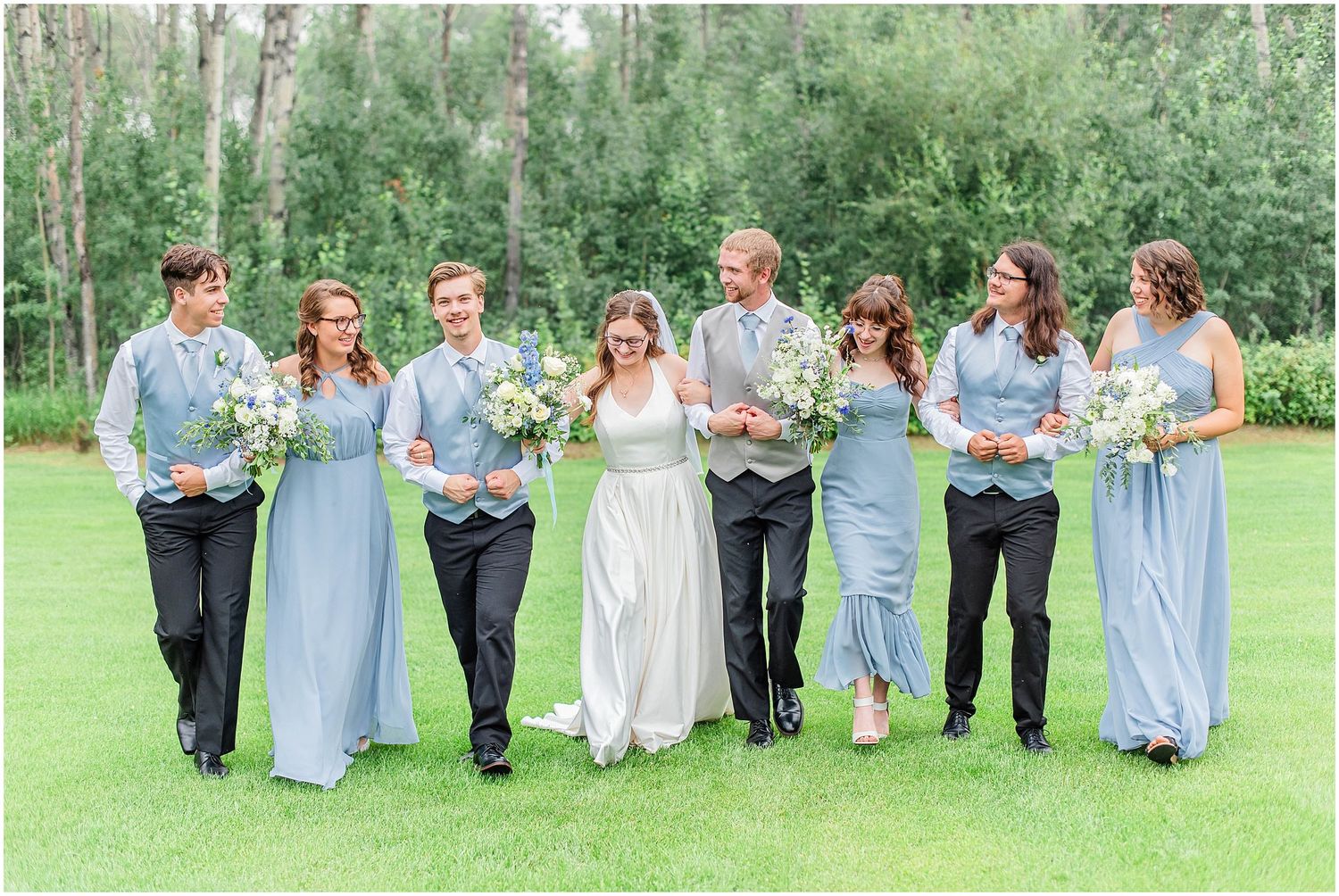 Wedding party walking together on grass, bridesmaids in blue dresses and groomsmen in black suits.