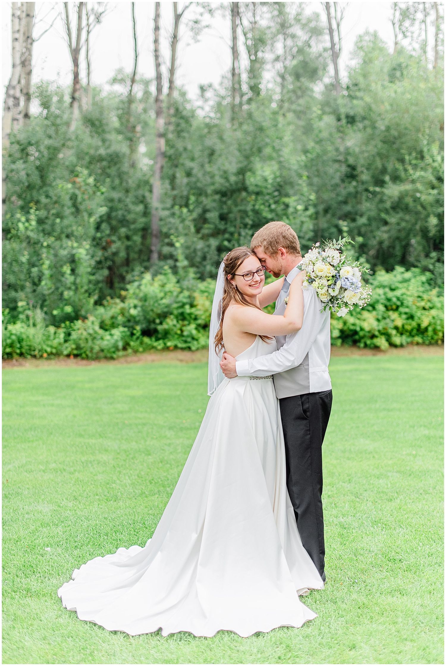 Bride and groom embracing tenderly on a lush green lawn surrounded by trees on their wedding day.
