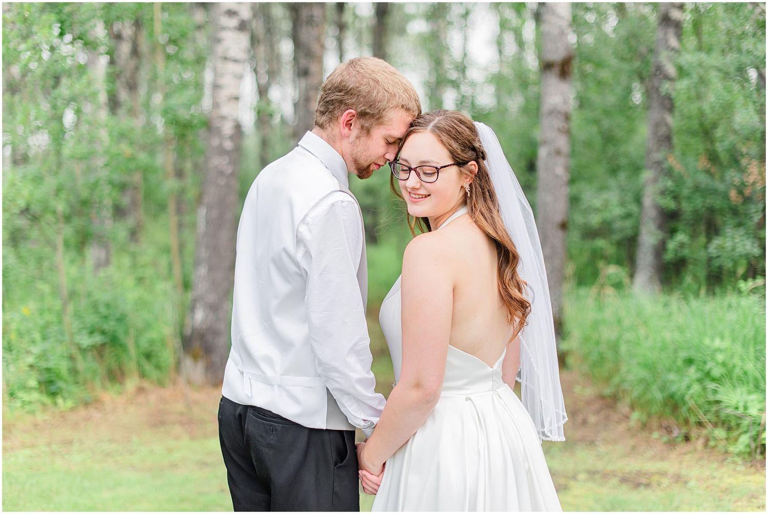 Bride and groom holding hands and smiling at each other in a wooded outdoor setting.