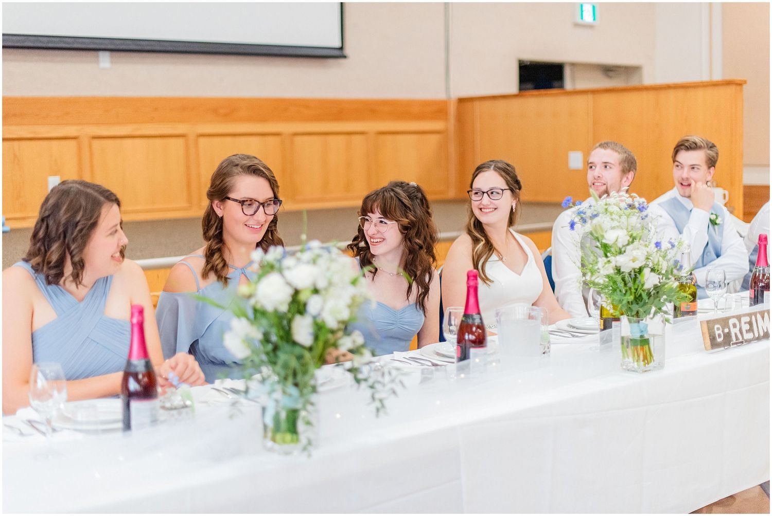 Wedding party seated at decorated head table laughing during reception celebration.