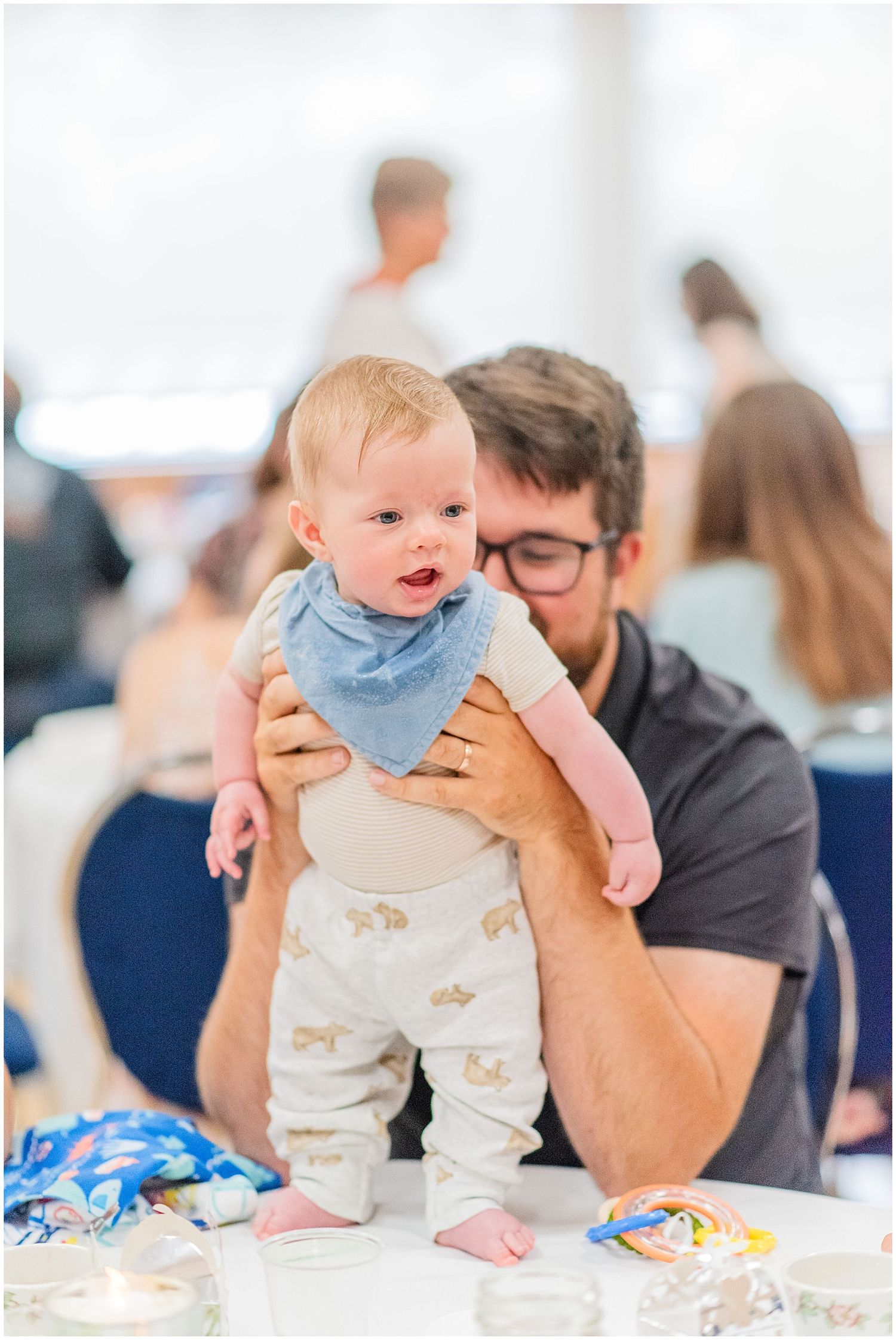 Father lovingly holds smiling baby dressed in blue bib and animal print pants at wedding reception.