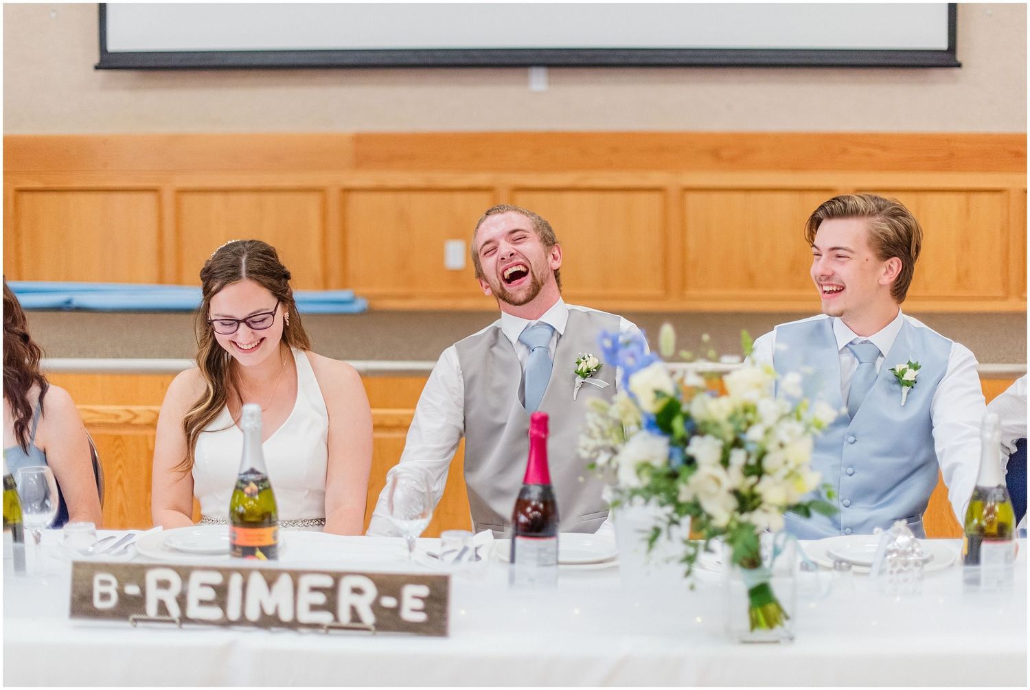 Bride and groom laughing joyfully at the wedding reception head table with guests nearby.