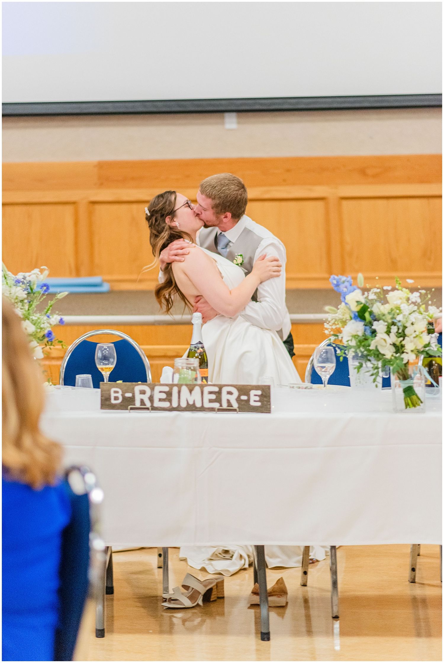 Bride and groom share a kiss at their wedding reception sweetheart table decorated with blue floral arrangements.