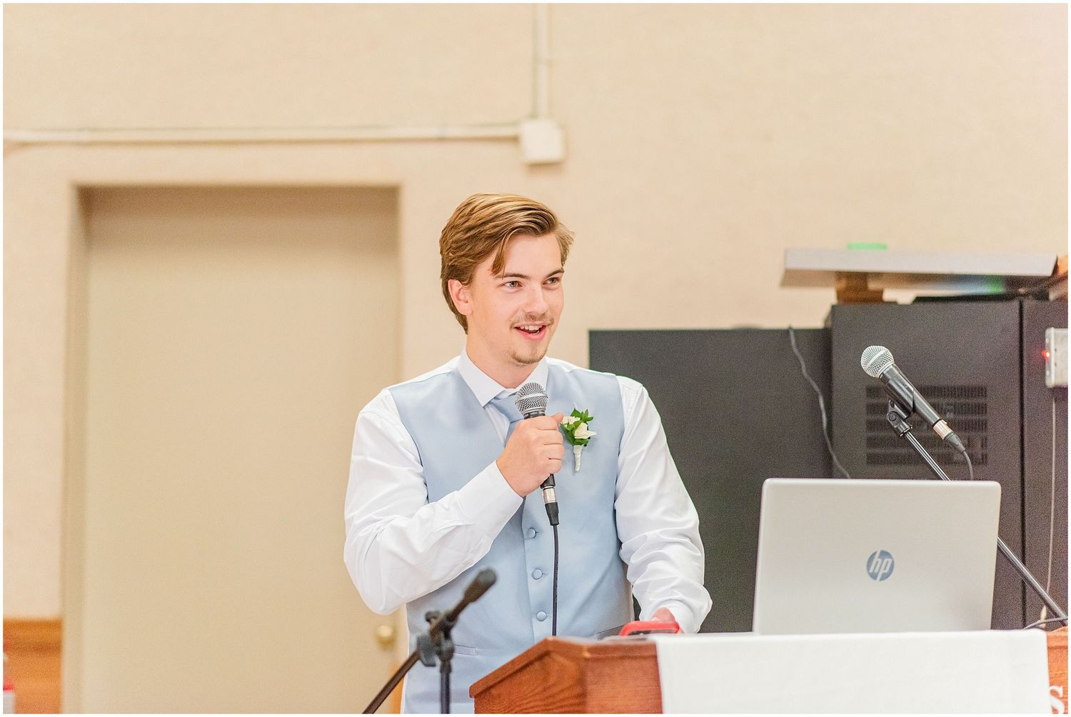 Smiling groomsman in light blue vest delivers speech at wedding reception podium with microphone.