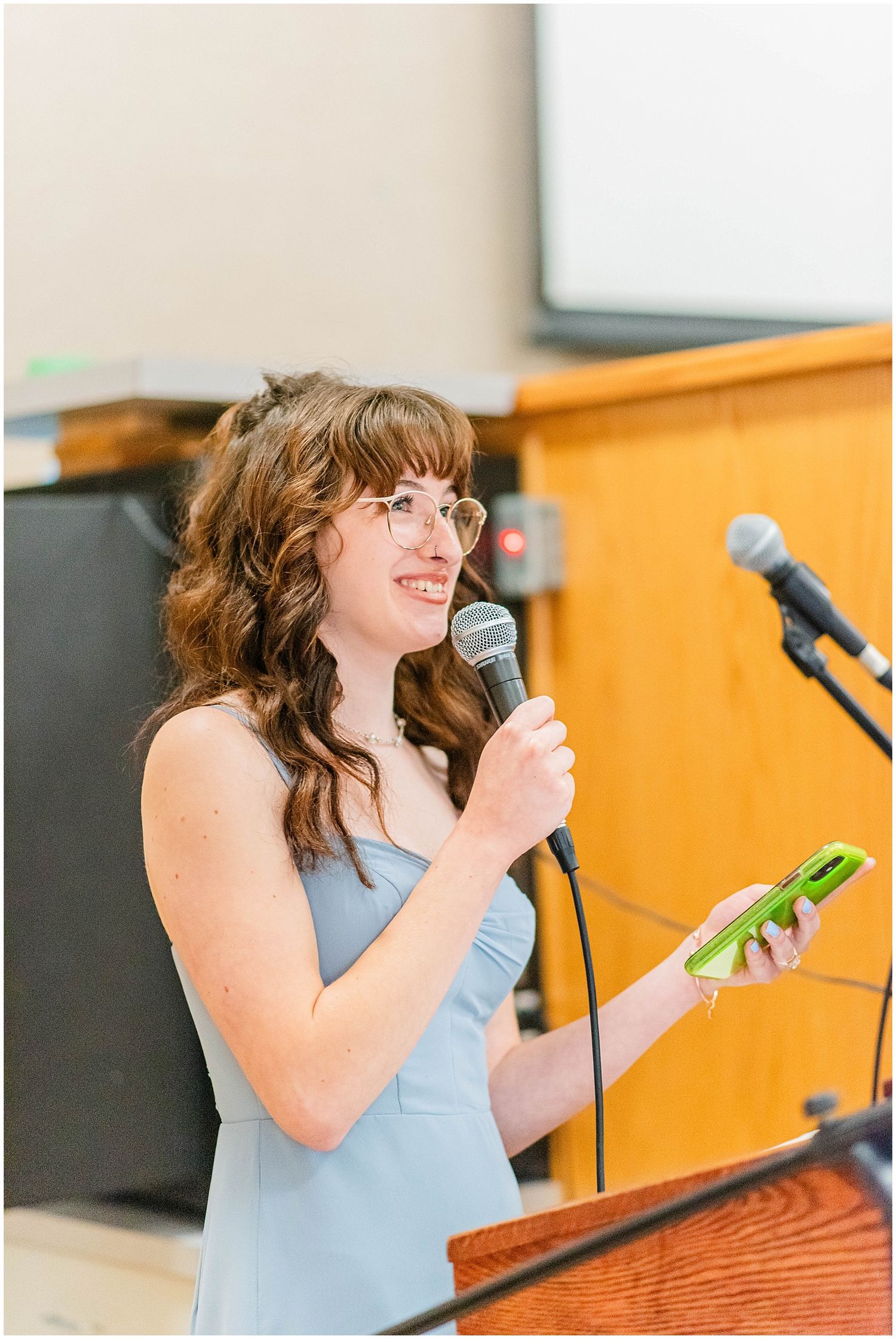 Bridesmaid in blue dress giving speech at wedding reception podium with microphone.