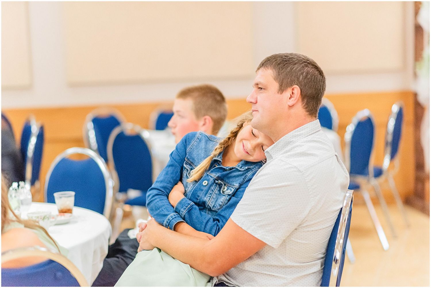 Young girl rests her head on a mans shoulder while seated at a wedding reception table.