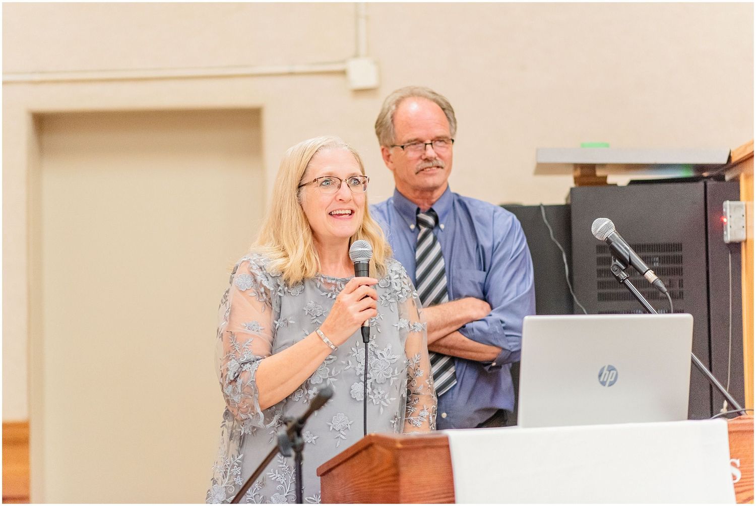 Woman speaking at podium with microphone while man stands behind her at indoor event.