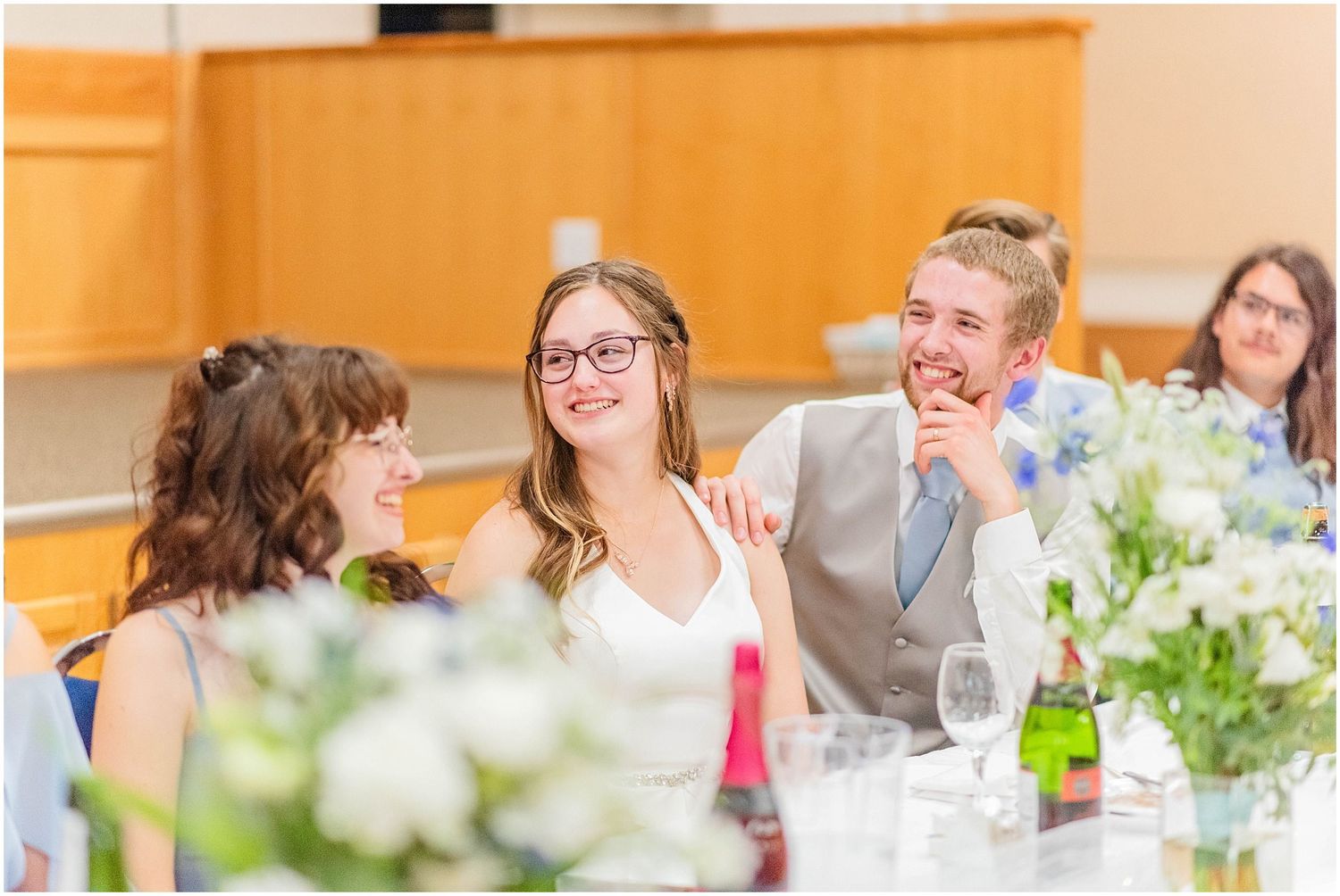 Bride and groom laugh joyfully with guests while seated at their wedding reception dinner table.