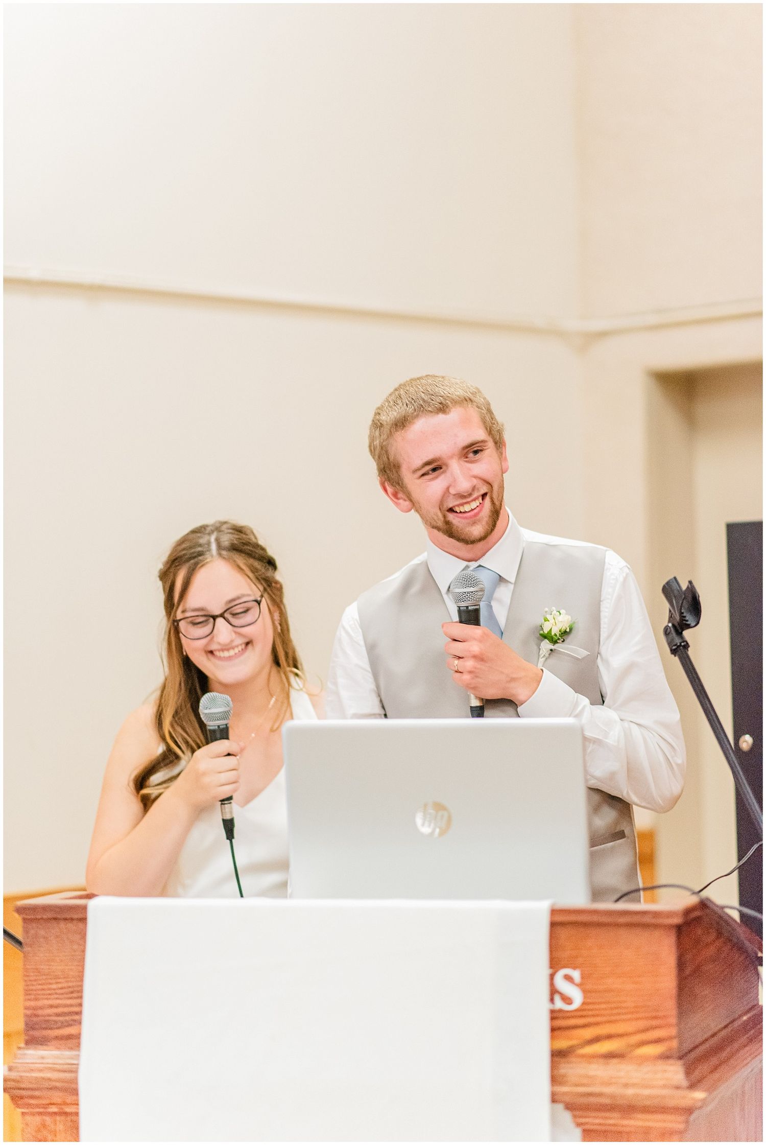 Smiling bride and groom speaking at a podium with microphones during their wedding reception.