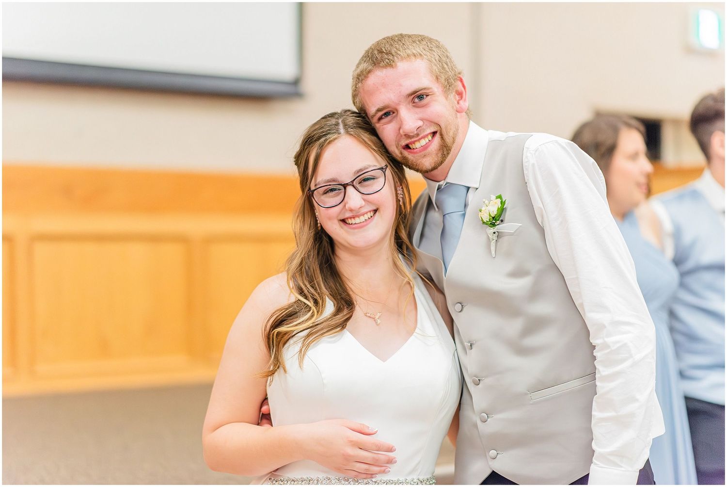 Smiling bride and groom pose together at their wedding reception, bride wearing glasses and white gown, groom in gray vest.
