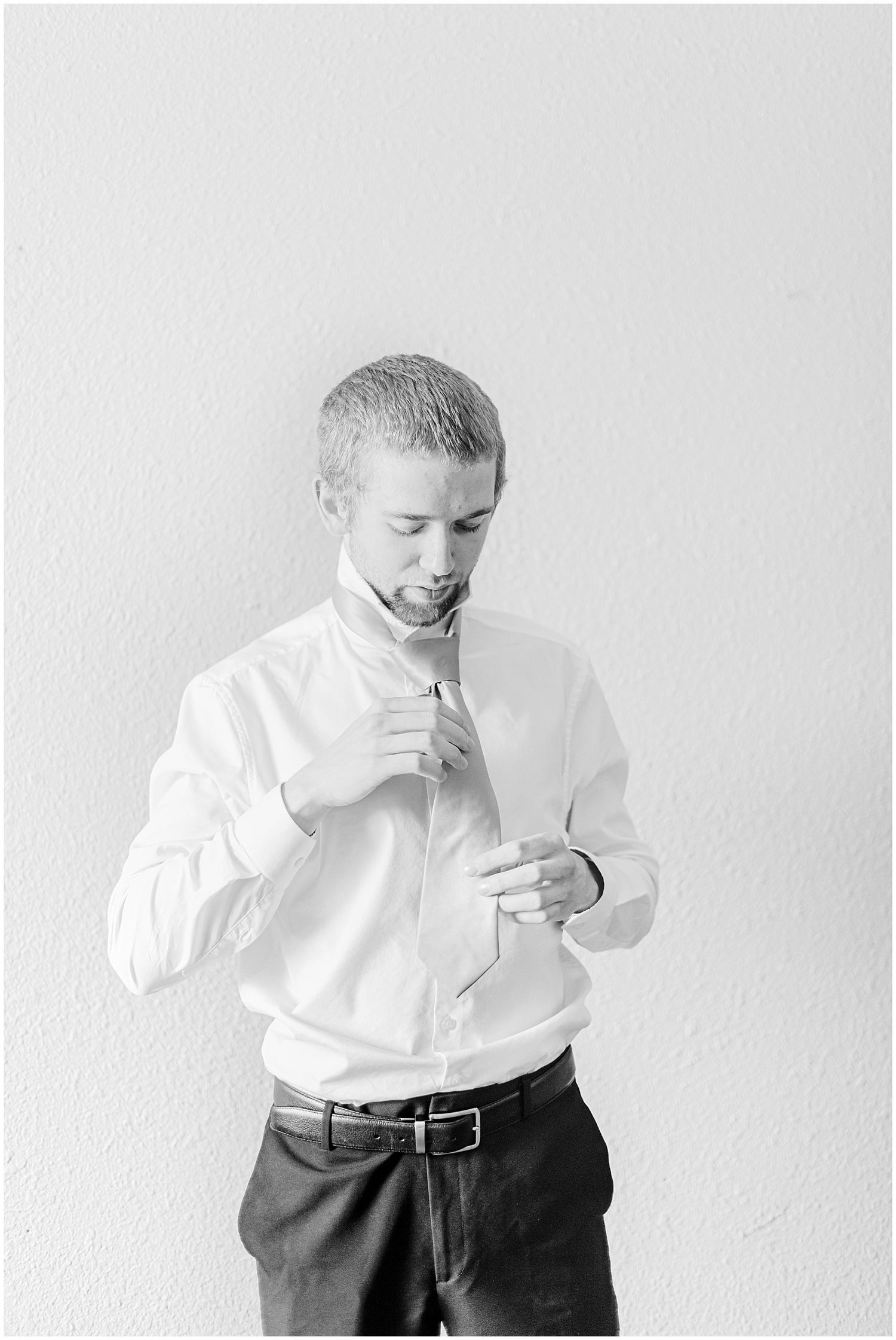 Groom in white dress shirt adjusts his tie while getting ready, captured in classic black and white.