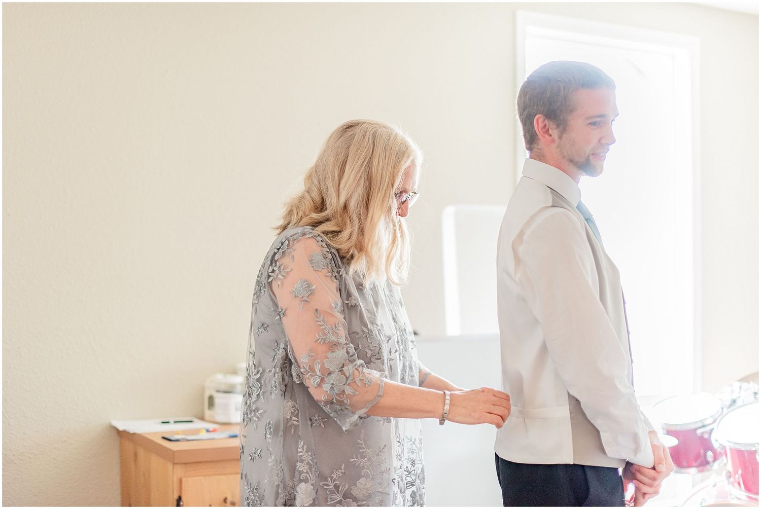 A woman in a gray lace dress helps a groom button his shirt before the wedding ceremony.