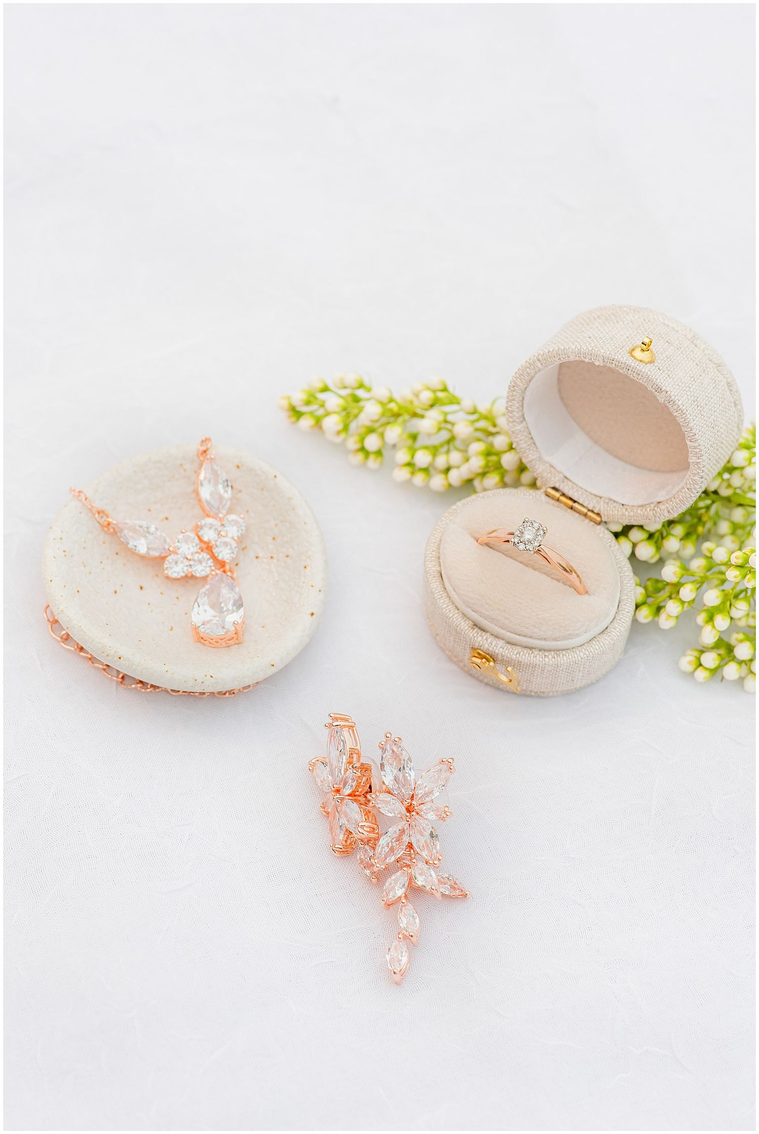 Rose gold engagement ring in velvet box beside crystal earrings on macarons with lily of the valley flowers.