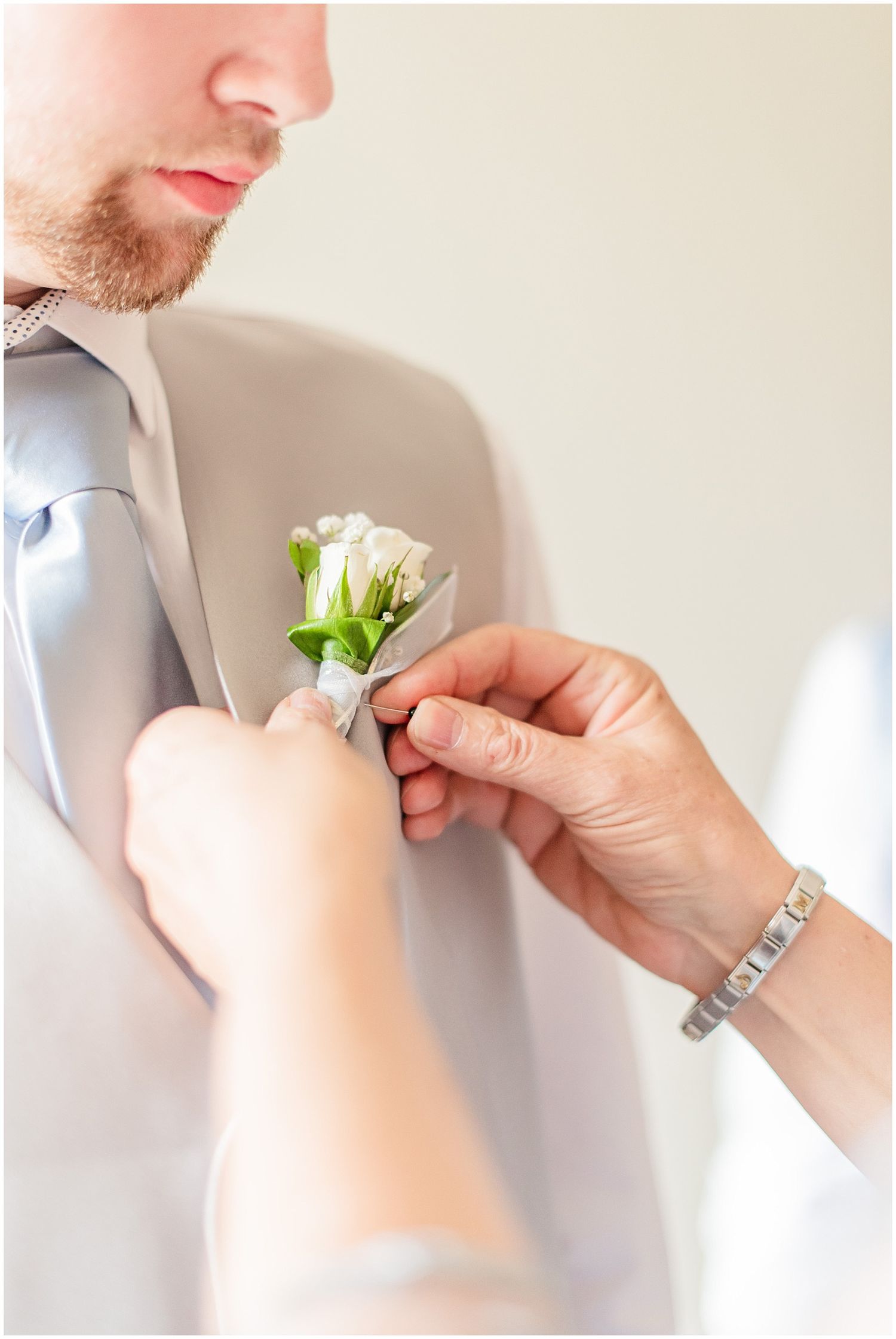 Someone pins a white rose boutonniere onto a groom wearing a gray vest and light blue tie.