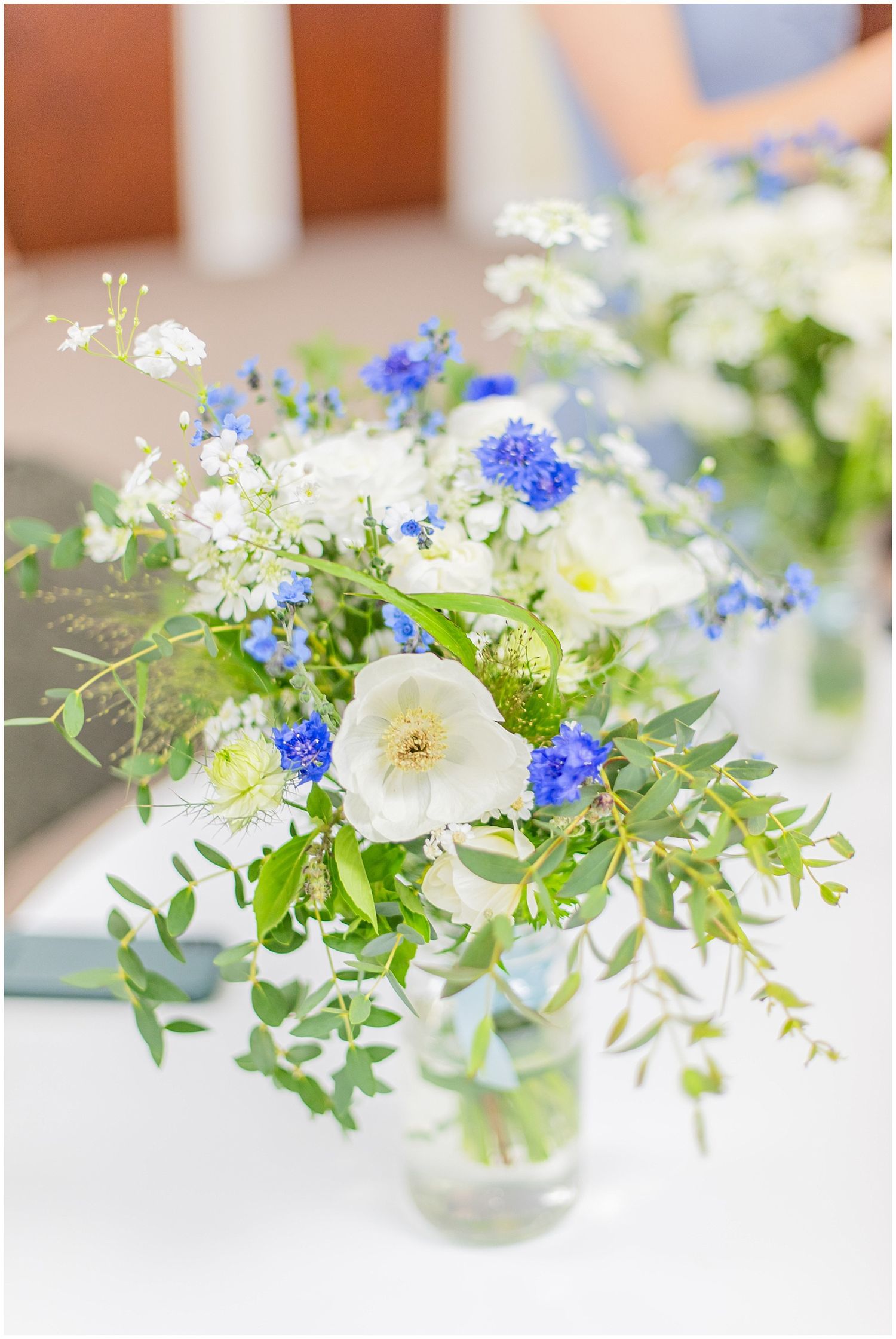 Blue and white wildflower wedding centerpiece in glass vase with greenery on white table.