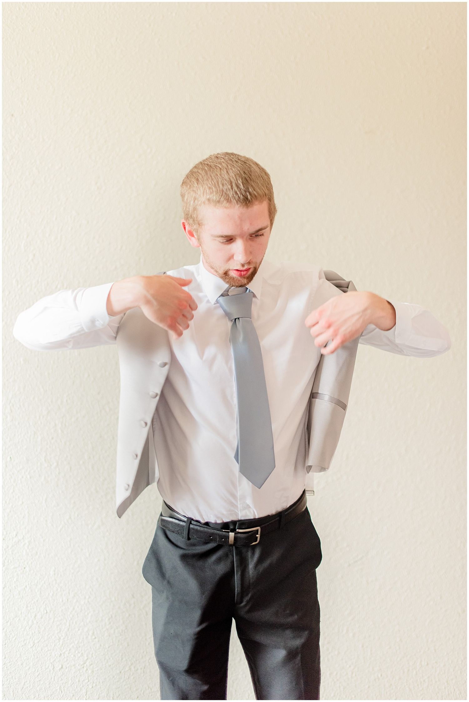 Groom in white shirt and grey tie putting on suit jacket before wedding ceremony.