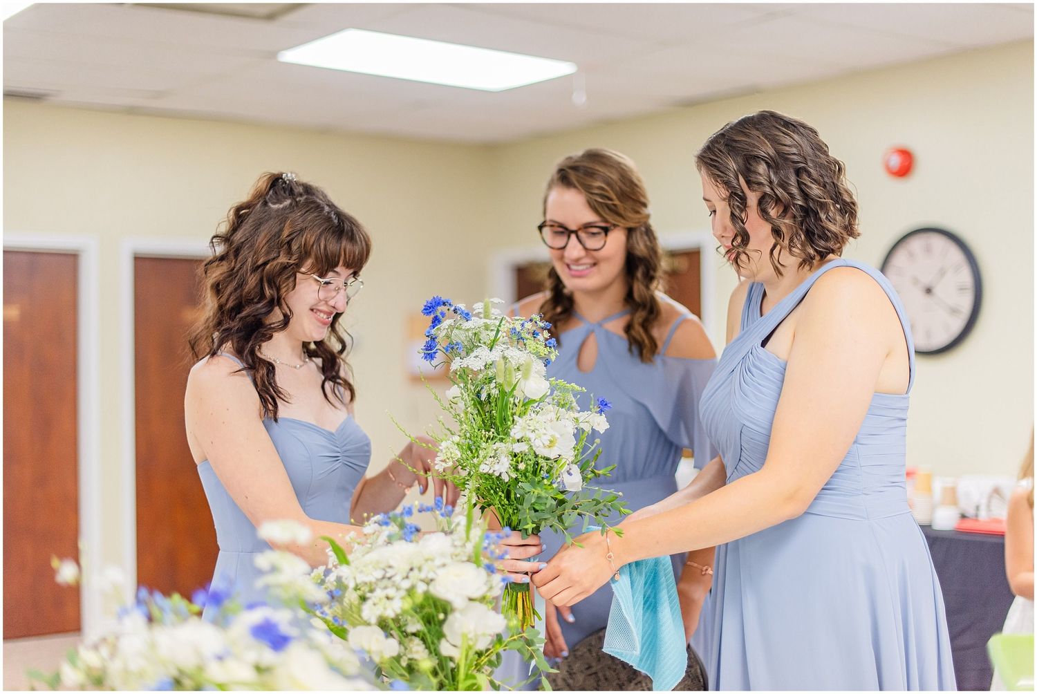 Three bridesmaids in blue dresses arranging wildflower bouquets together indoors before a wedding ceremony.