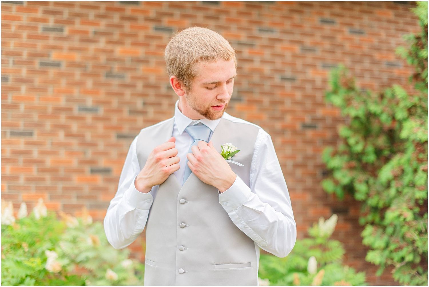 Groom in gray vest adjusting his boutonniere outdoors against a brick wall before wedding.