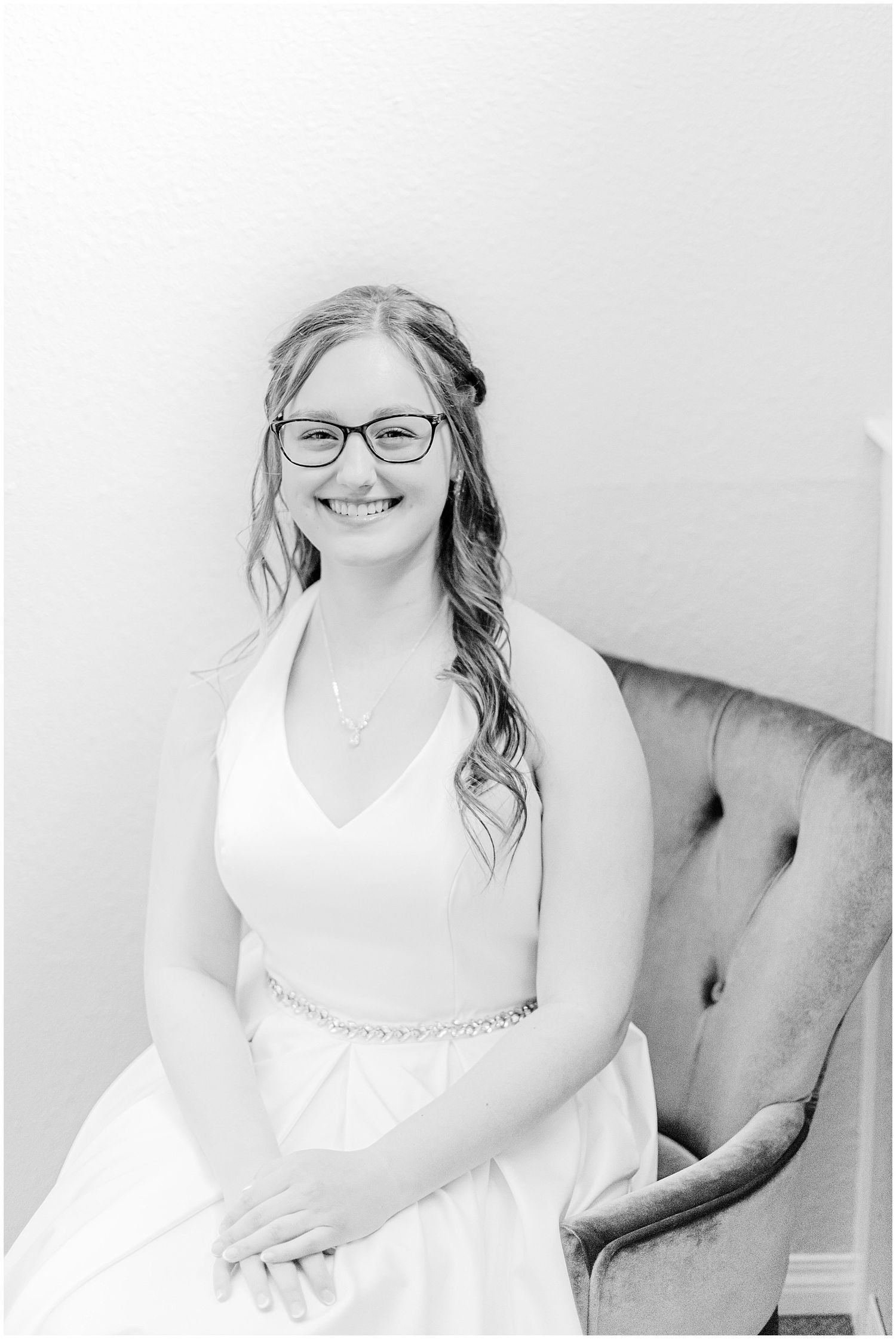 Smiling bride in white gown with glasses seated in velvet chair, elegant black and white portrait.