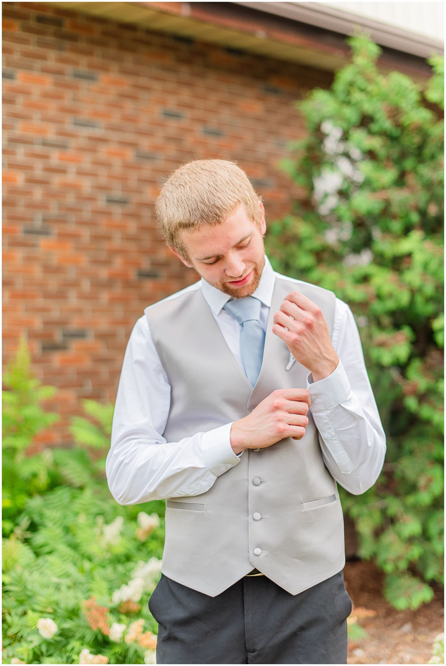 Smiling groom in gray vest and blue tie adjusts his cufflinks outside a brick building.
