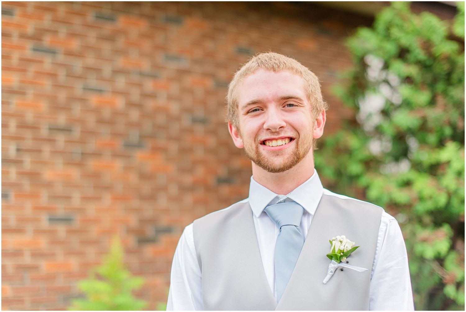 Smiling groom in a gray vest and blue tie with a boutonniere outside a brick building.
