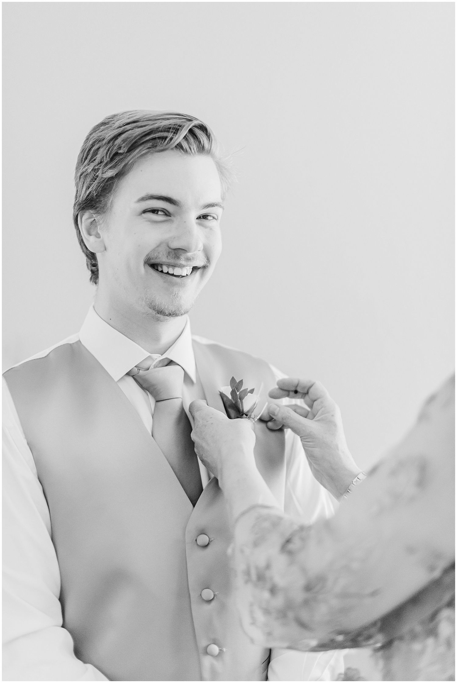 Smiling groom in vest and tie having boutonniere pinned on, black and white photo.