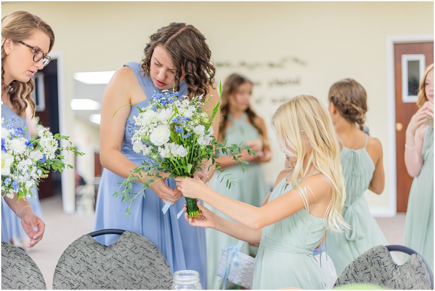 Bridesmaids in blue and sage dresses arranging white and blue floral bouquets before a wedding ceremony.