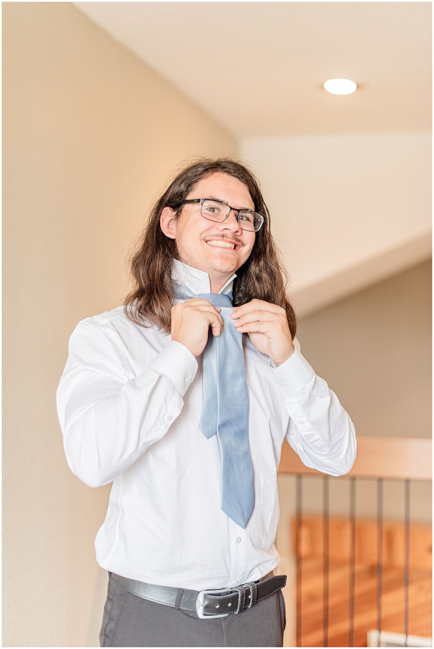 Smiling groom with long hair adjusts his light blue tie while getting ready indoors.