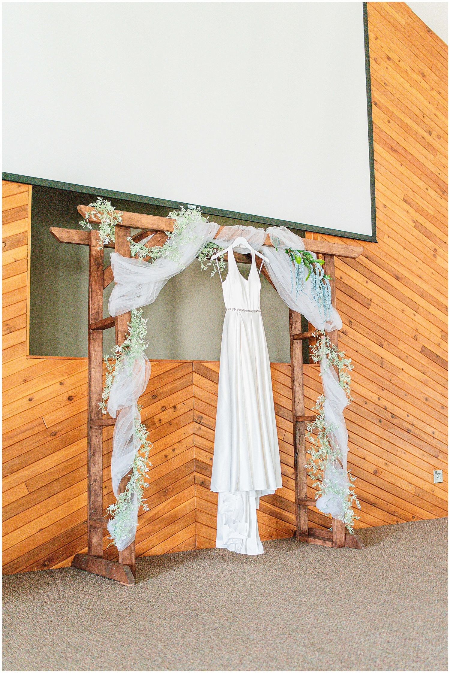 White wedding dress hanging on a rustic wooden arch decorated with greenery and sheer fabric in a wood-paneled venue.
