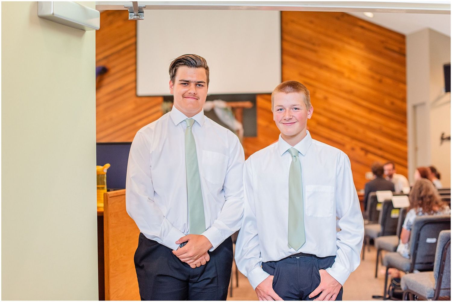 Two young groomsmen in white shirts and sage green ties smiling together inside a wedding venue.