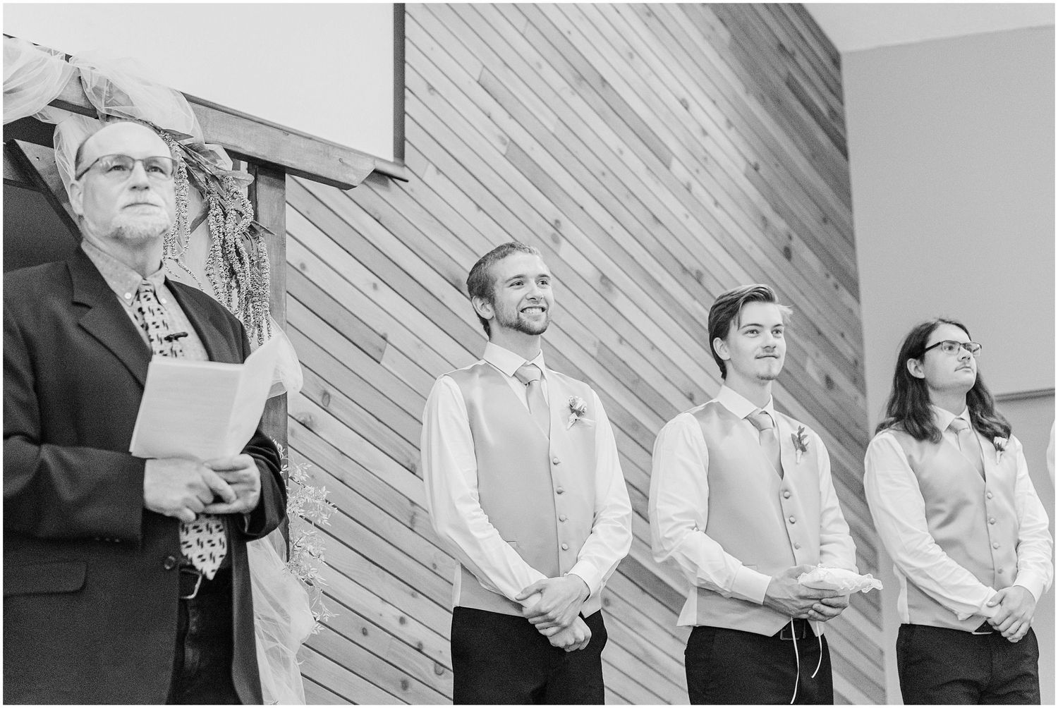 Black and white photo of groomsmen standing at wedding altar in vests and ties during ceremony.