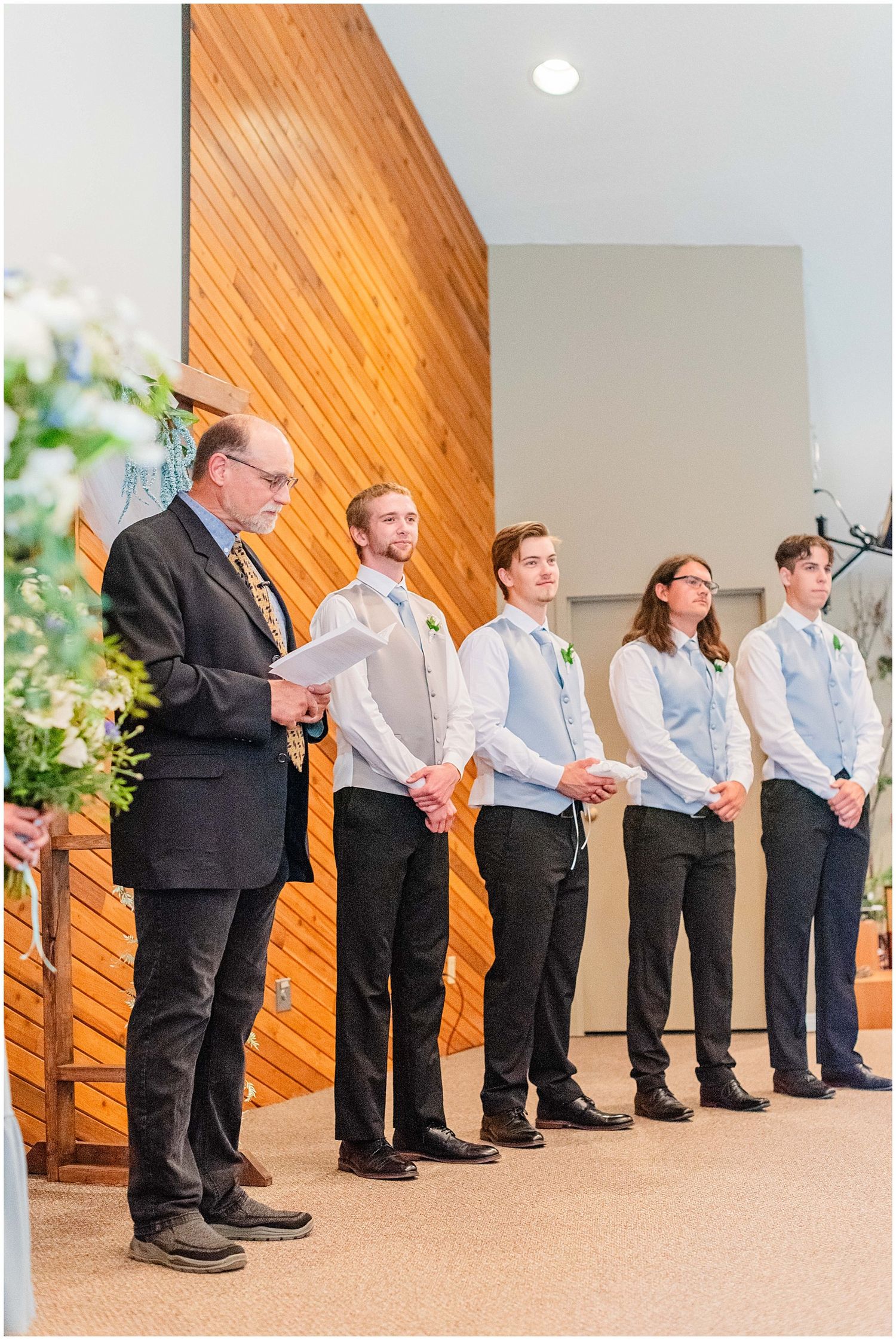 Groomsmen in gray vests and dark pants stand at the altar with an officiant holding papers during a wedding ceremony.