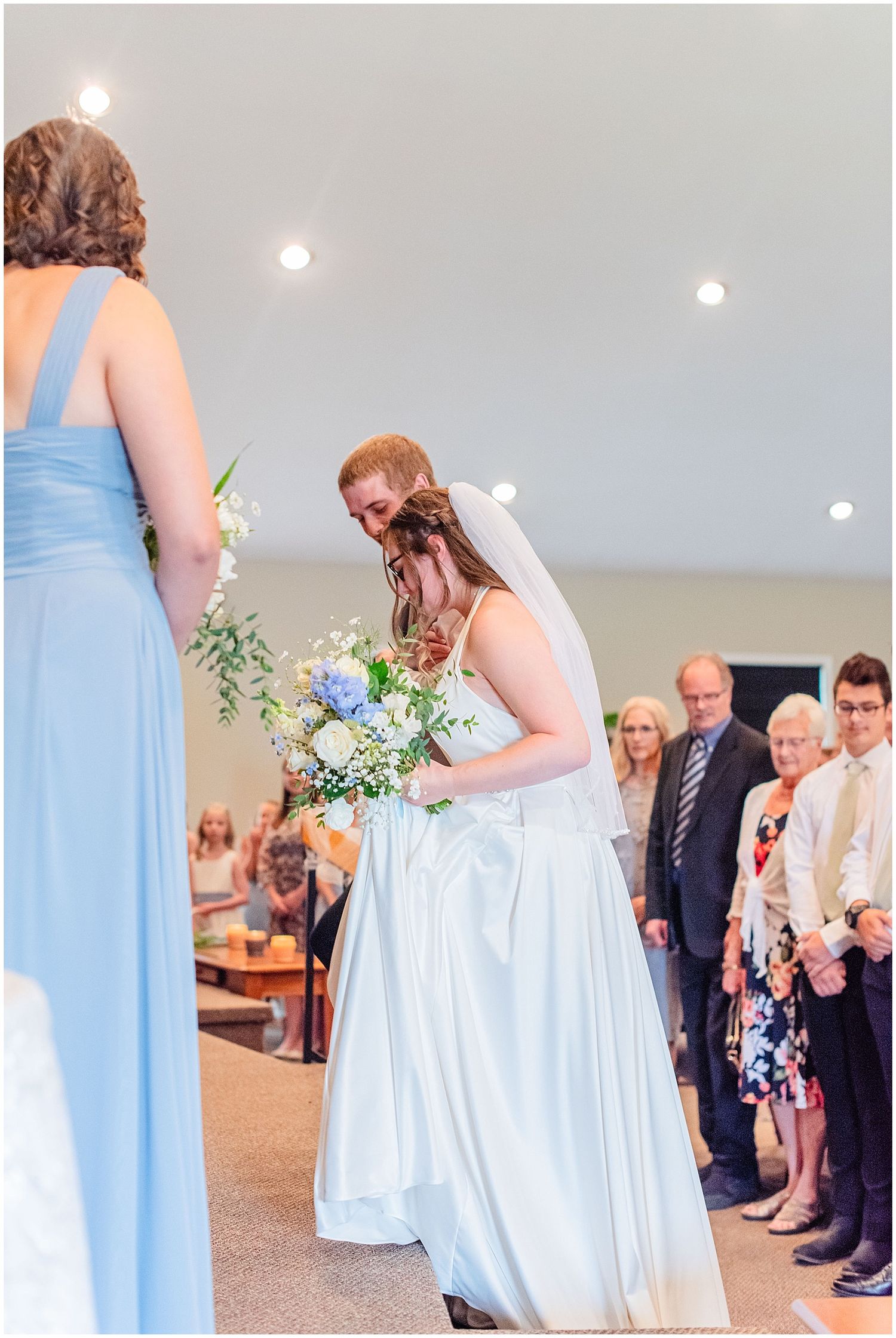 Bride in white gown bows to greet bridesmaid in blue dress while holding floral bouquet during wedding ceremony.