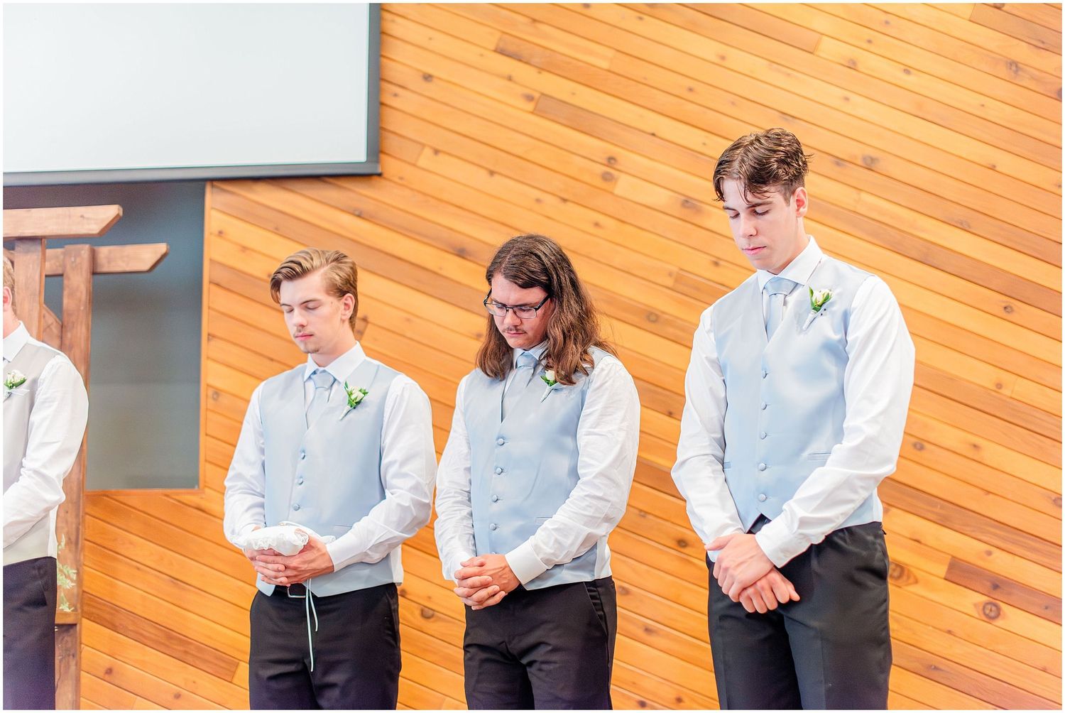 Three groomsmen in light blue vests and white shirts standing solemnly during a wedding ceremony.