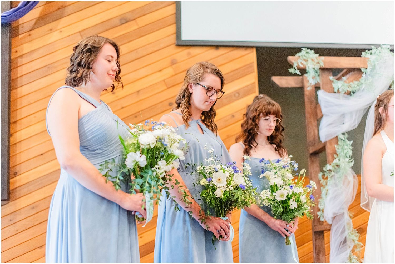 Three bridesmaids in light blue dresses holding white floral bouquets during wedding ceremony.