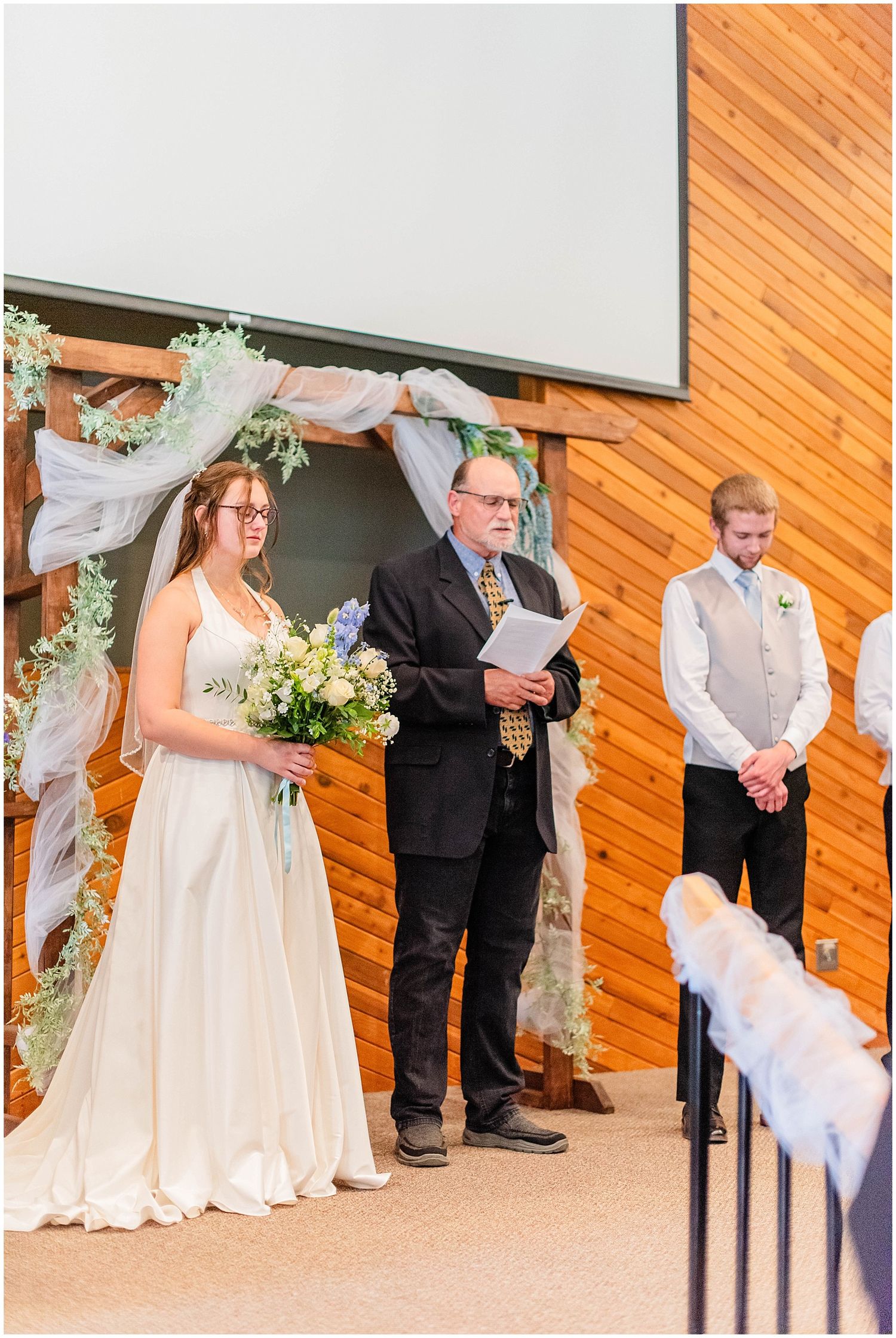 Bride in white gown holds bouquet beside officiant at decorated wooden arch during indoor wedding ceremony.