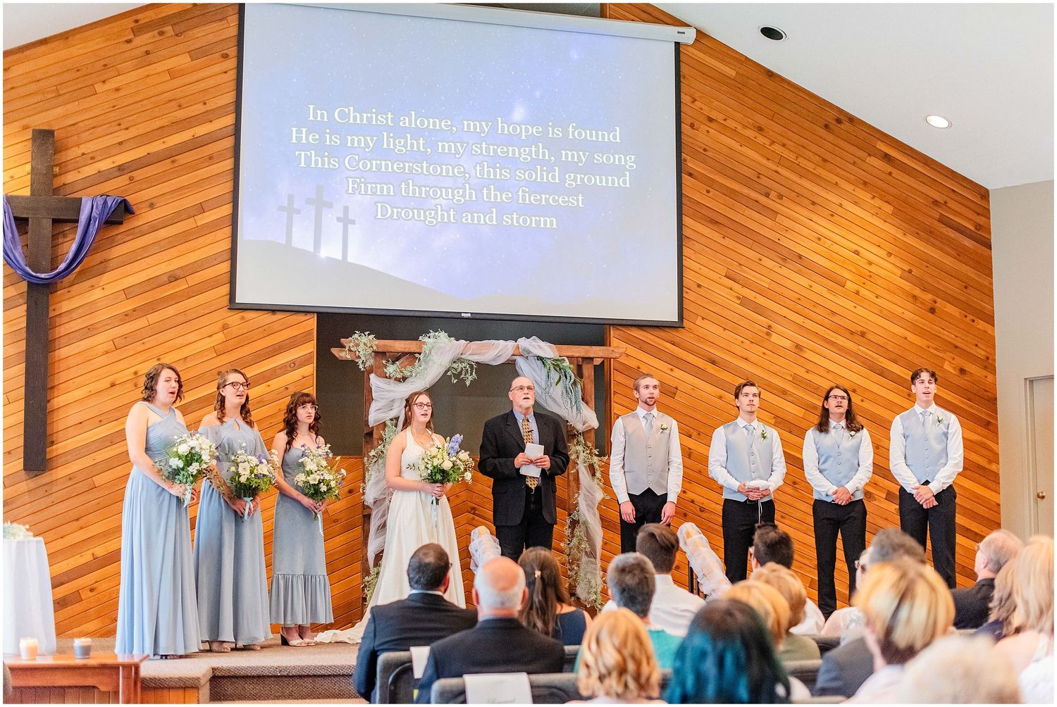 Wedding party stands at church altar with hymn lyrics projected on screen behind them.