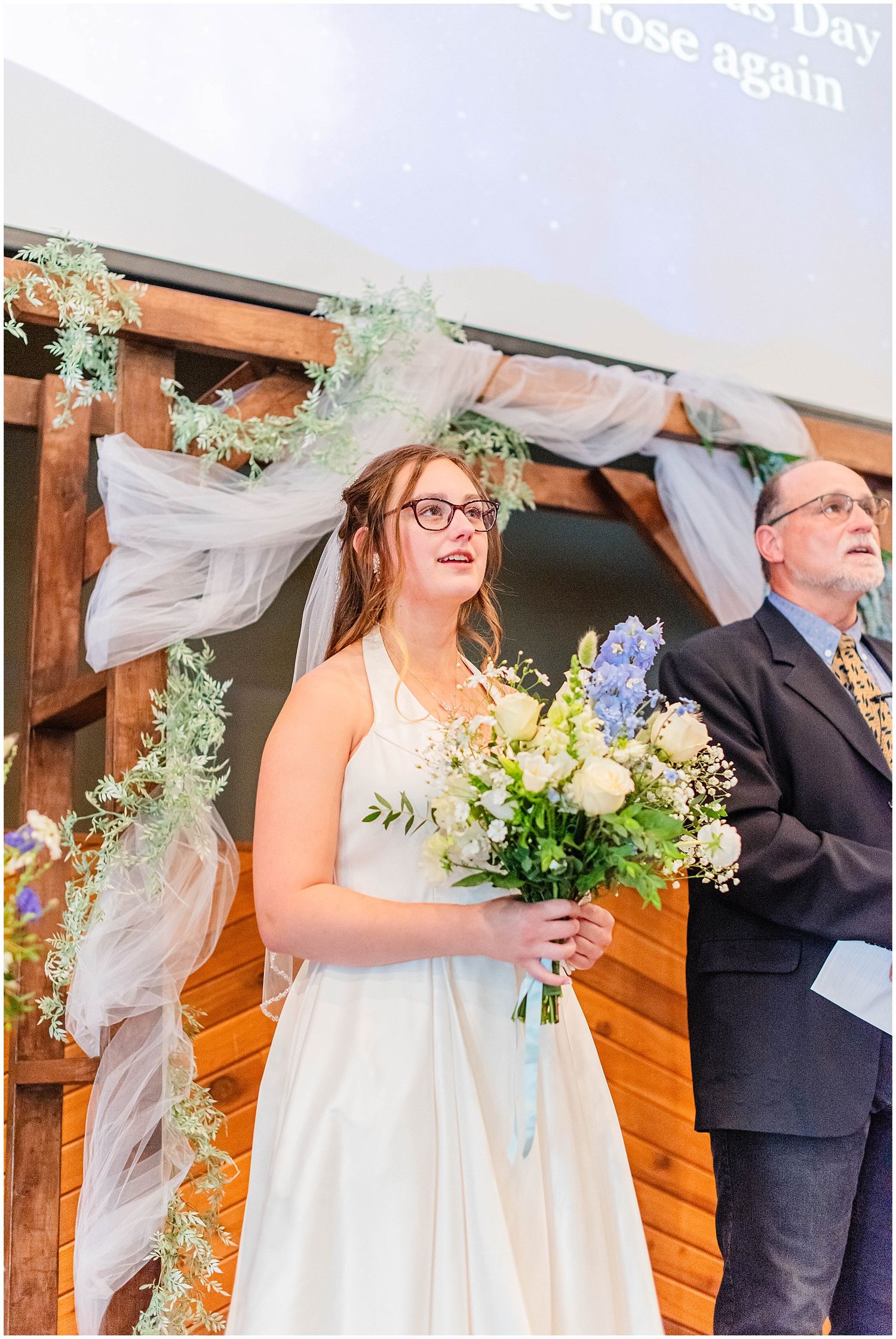 Bride in white gown holding blue and white bouquet standing at rustic wooden wedding altar.