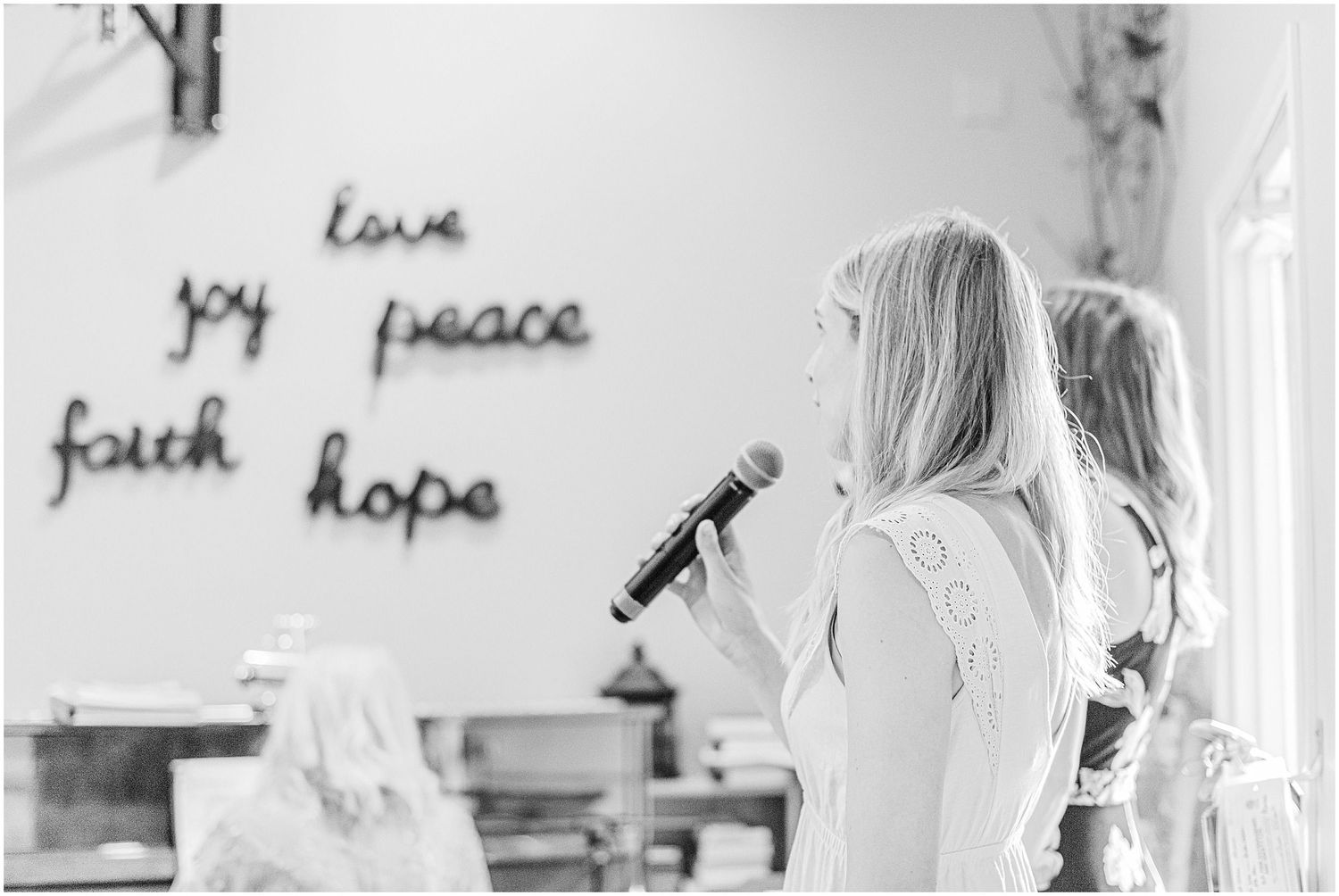 Blonde woman speaking into microphone at elegant event with love, joy, peace, faith, hope wall decor.