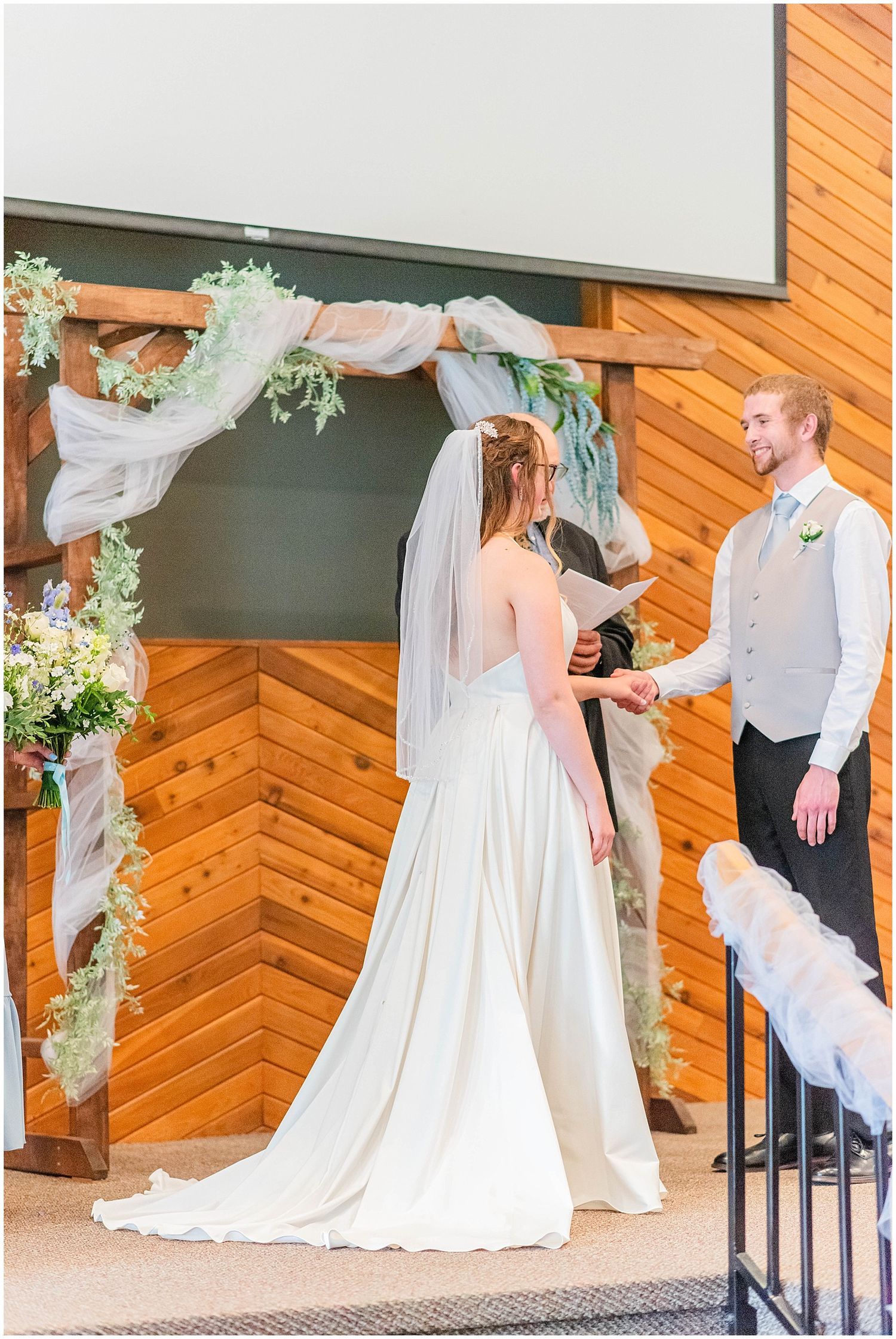 Bride and groom exchange vows under a rustic wooden arch adorned with greenery and sheer draping.