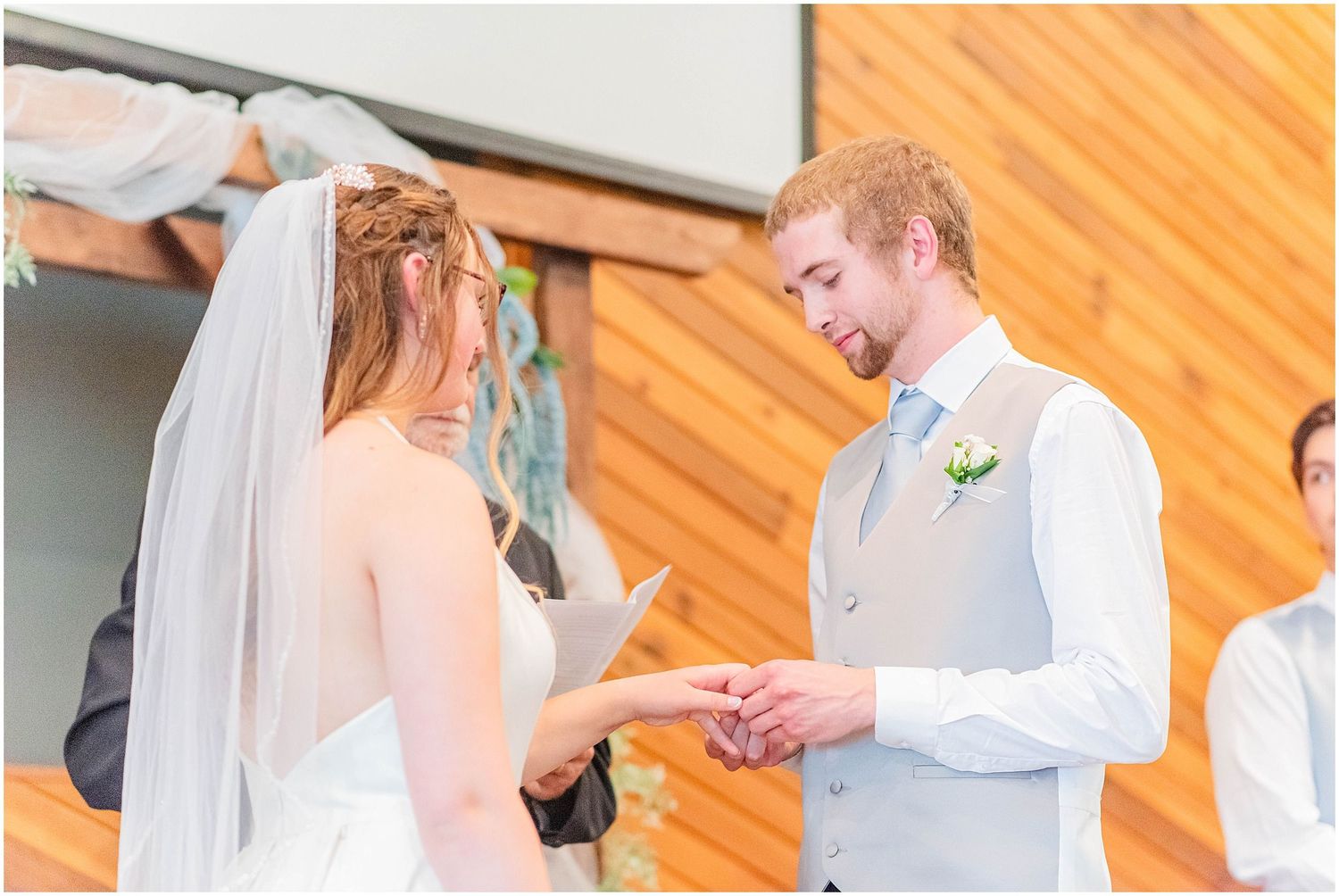 A groom in a gray vest places a ring on his brides finger during an outdoor wedding ceremony.