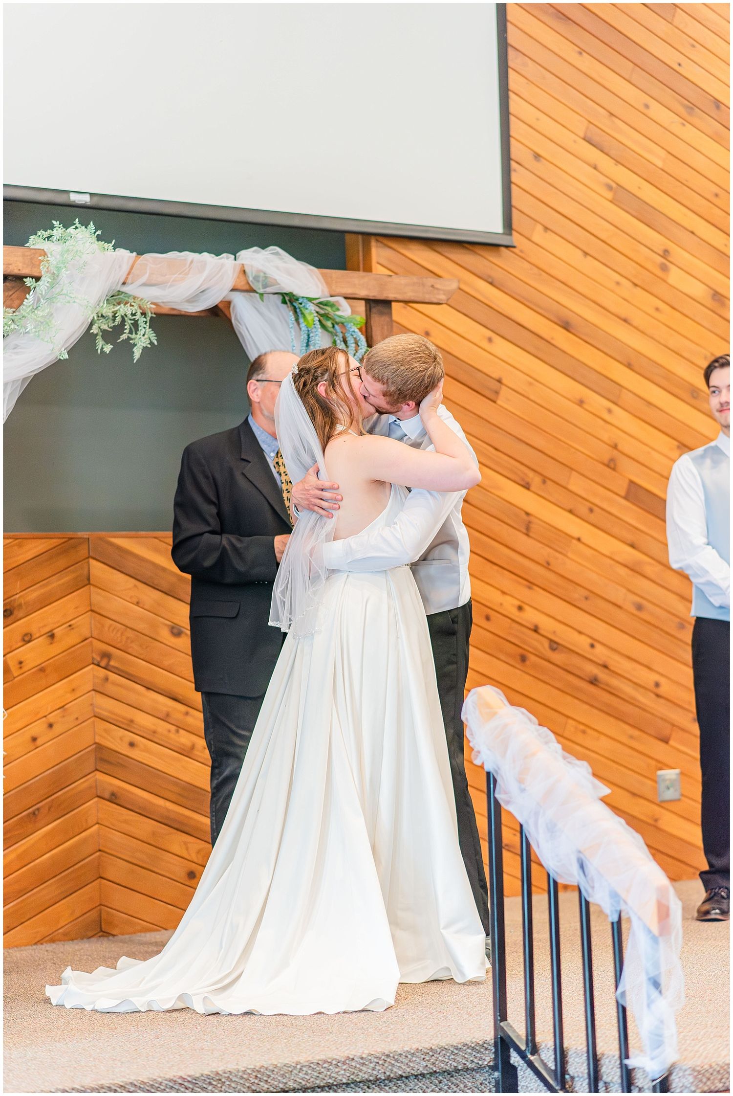 Bride and groom share first kiss at altar beneath floral arch in rustic wood-paneled church ceremony.