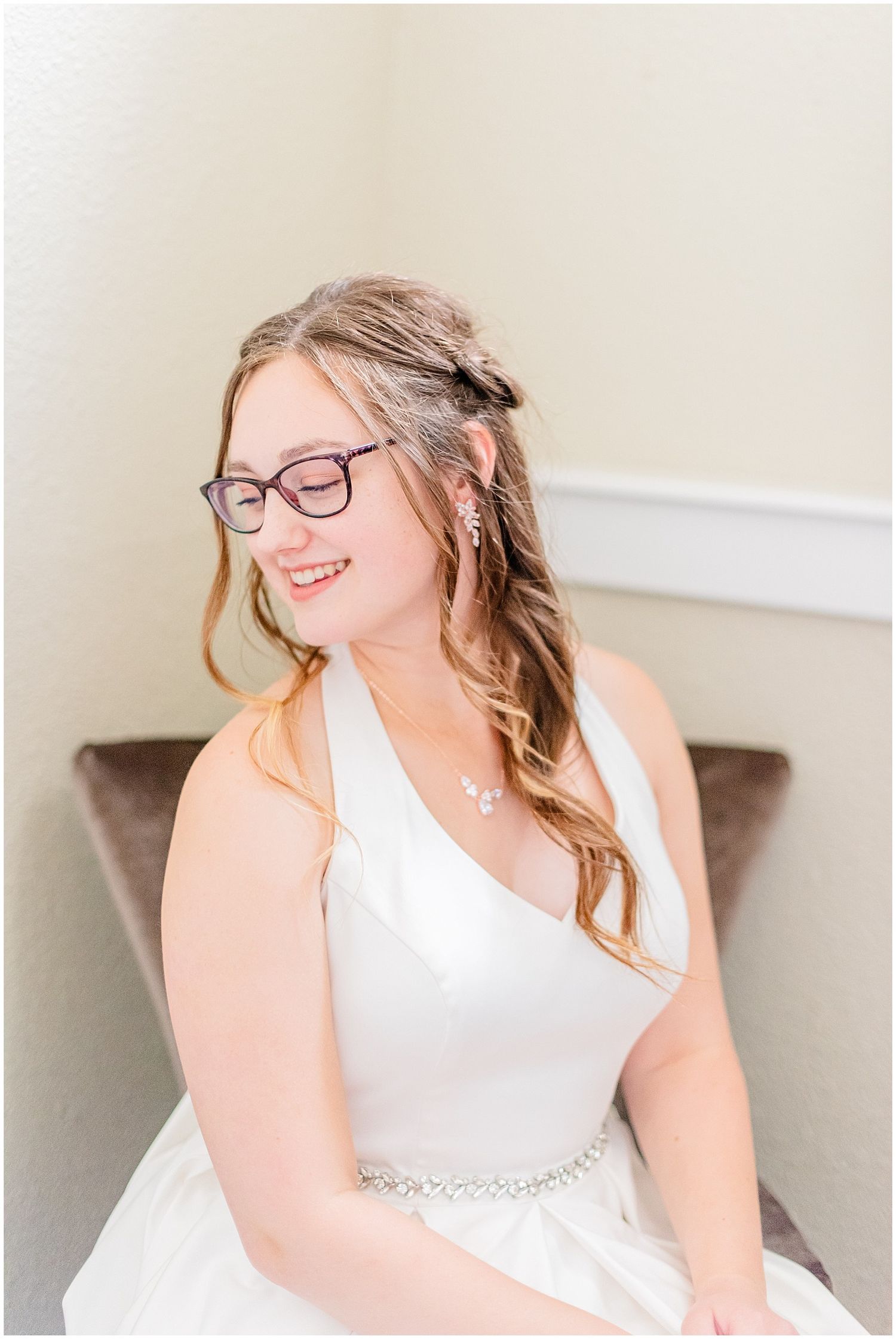 Smiling bride in white halter gown with embellished belt, wearing purple glasses and pearl jewelry, seated indoors.
