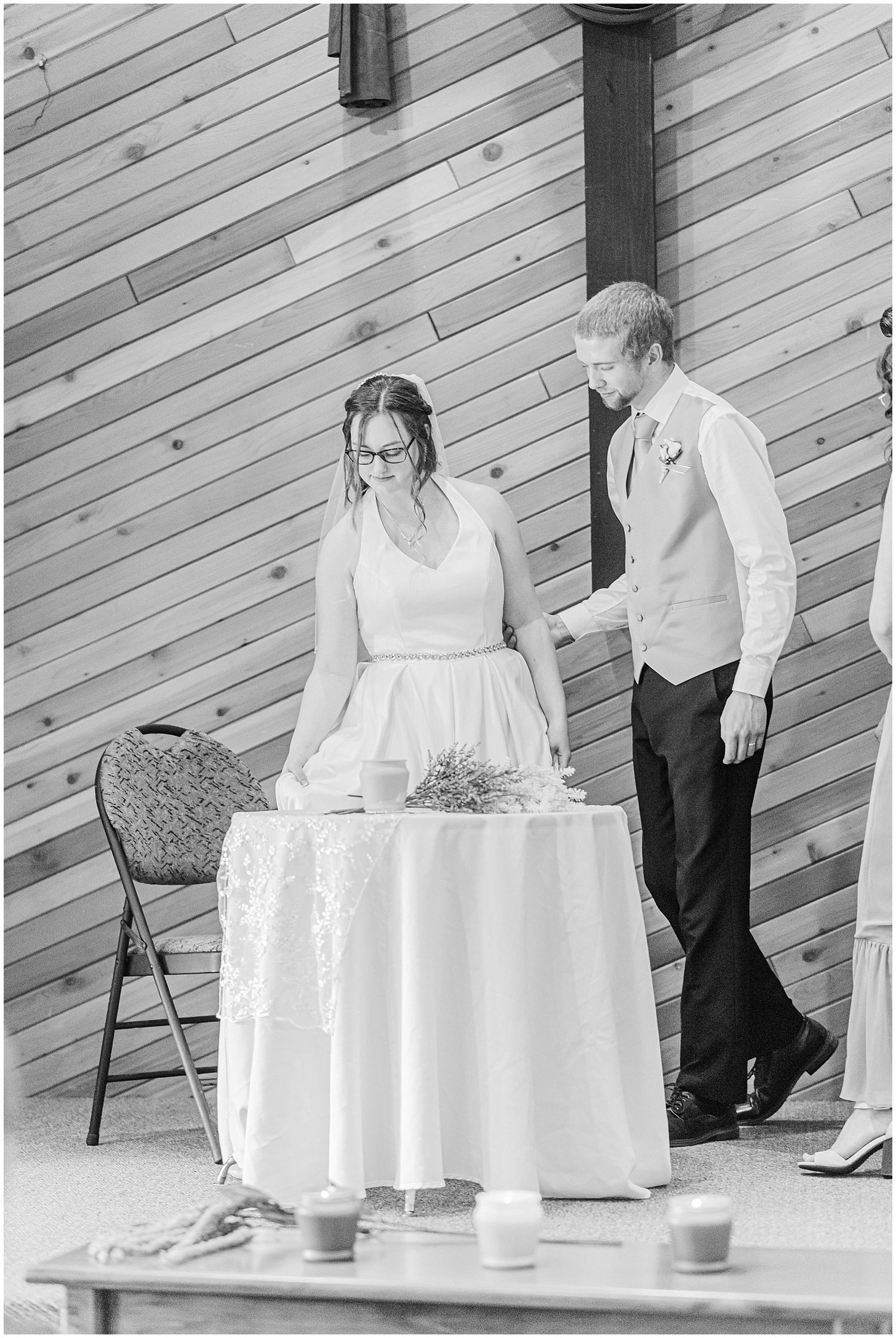 Bride and groom stand at a decorated table during their wedding reception in a rustic barn venue.