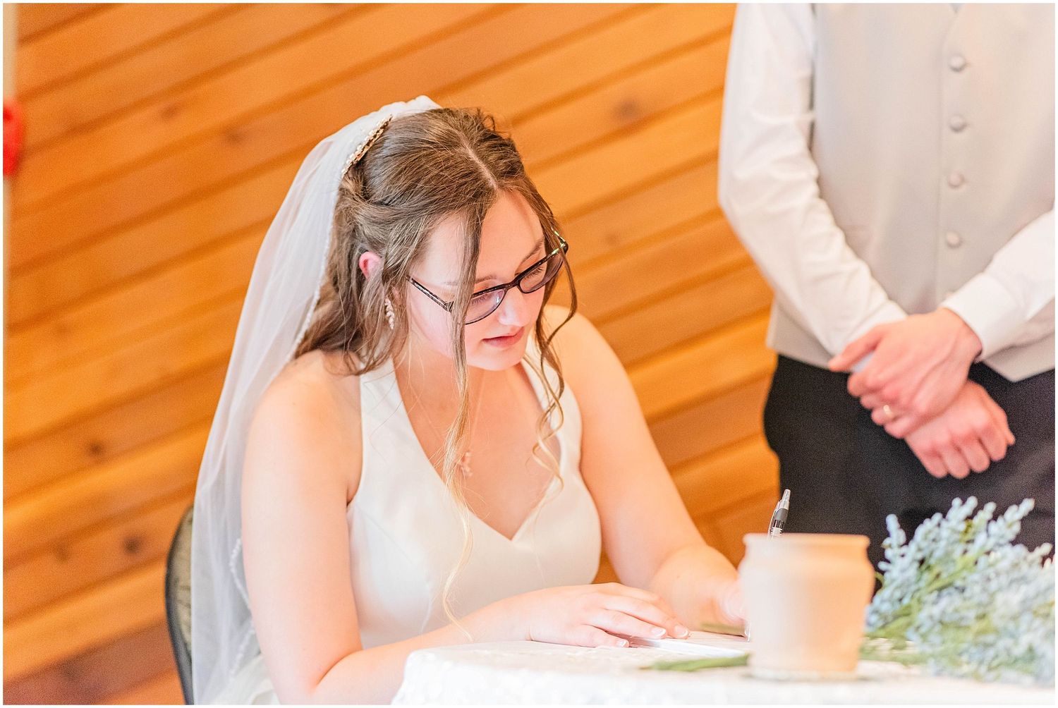 Bride wearing veil and glasses signing wedding certificate at table during ceremony.