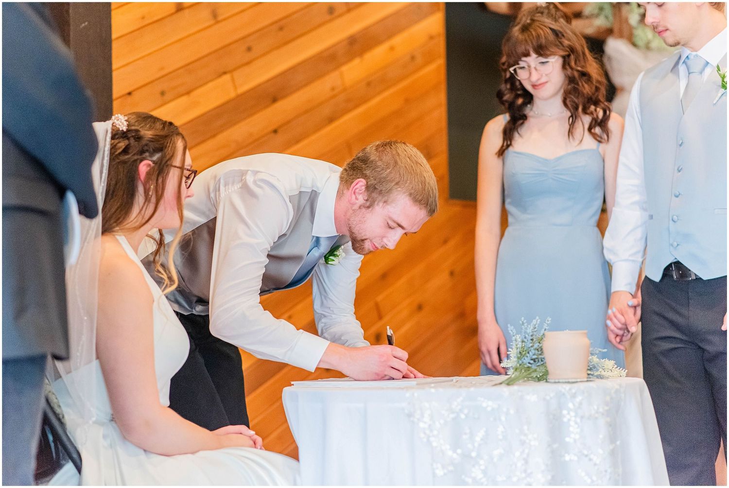Groom signing wedding documents at a white lace-covered table, flanked by bride and bridesmaid in blue dress.