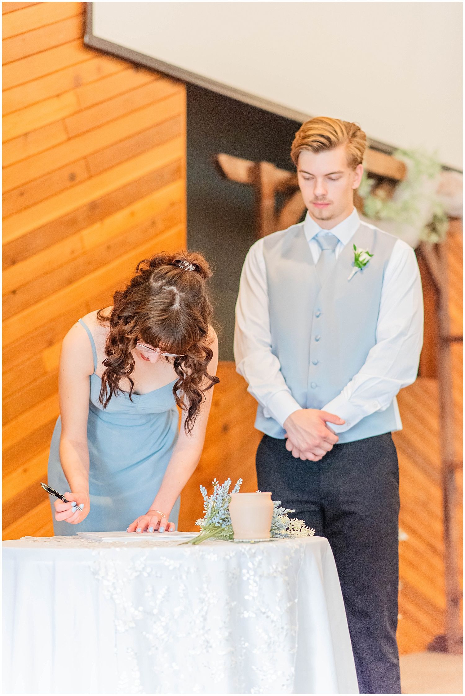 Bridesmaid in blue dress signing wedding documents while groom watches at decorated table.